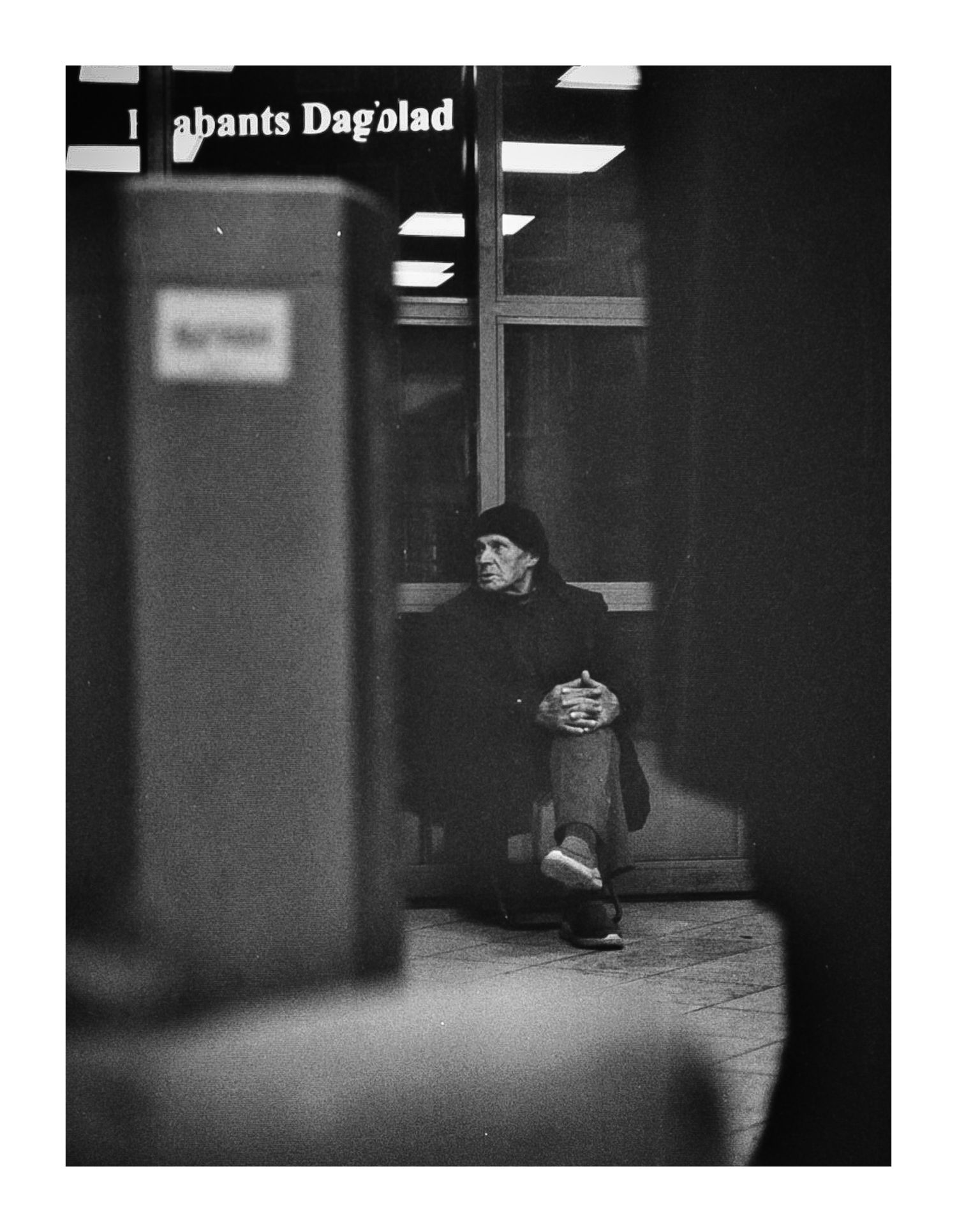 Elderly person sitting alone in dimly lit urban space, candid black and white photography.