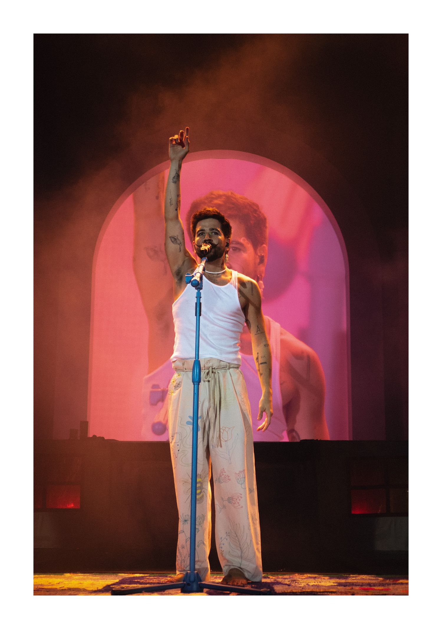performer on stage raising one hand under pink lights with an arch backdrop at a live concert.