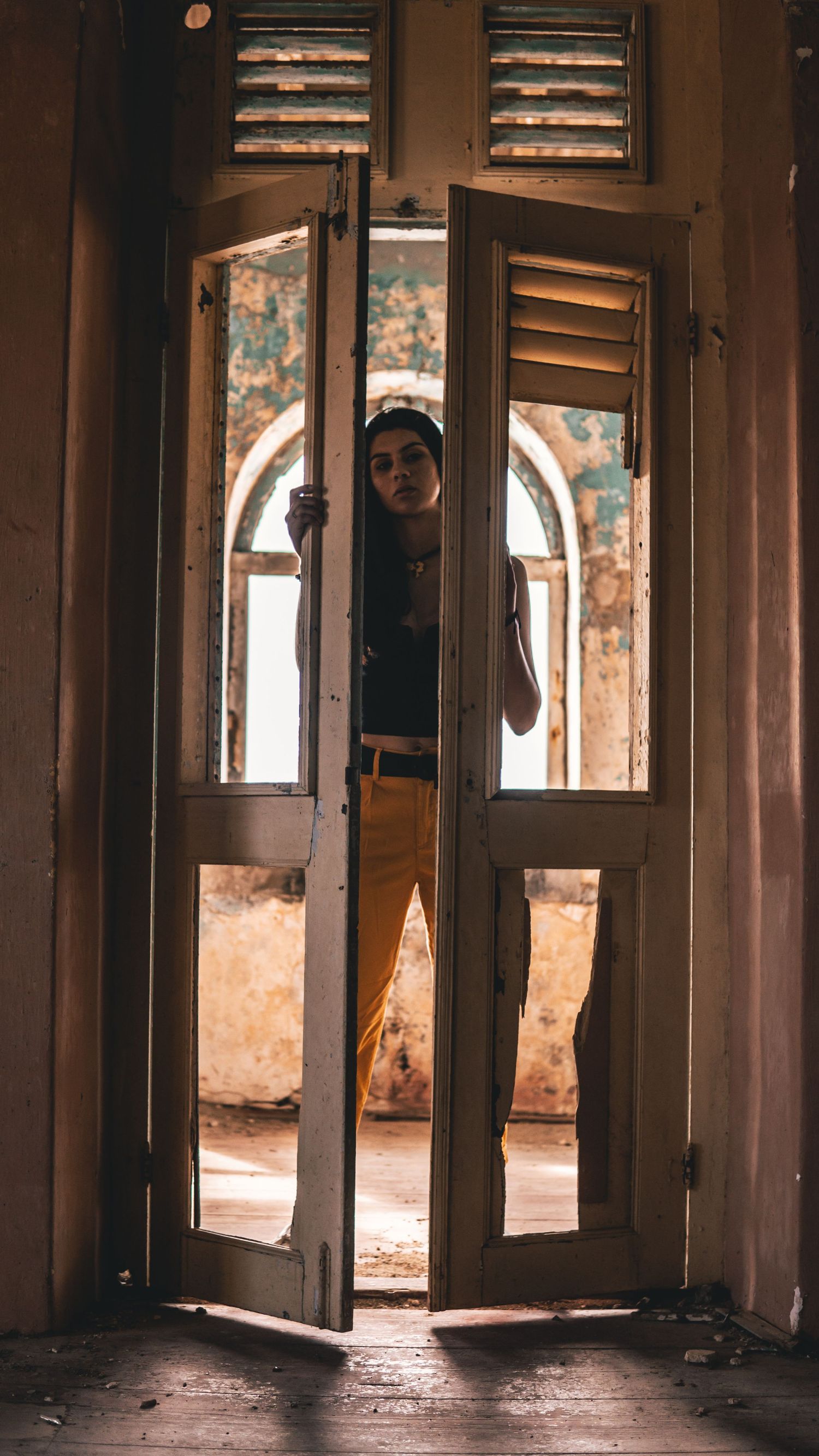 Woman standing in open vintage shuttered doors of a rustic abandoned building with arched windows.
