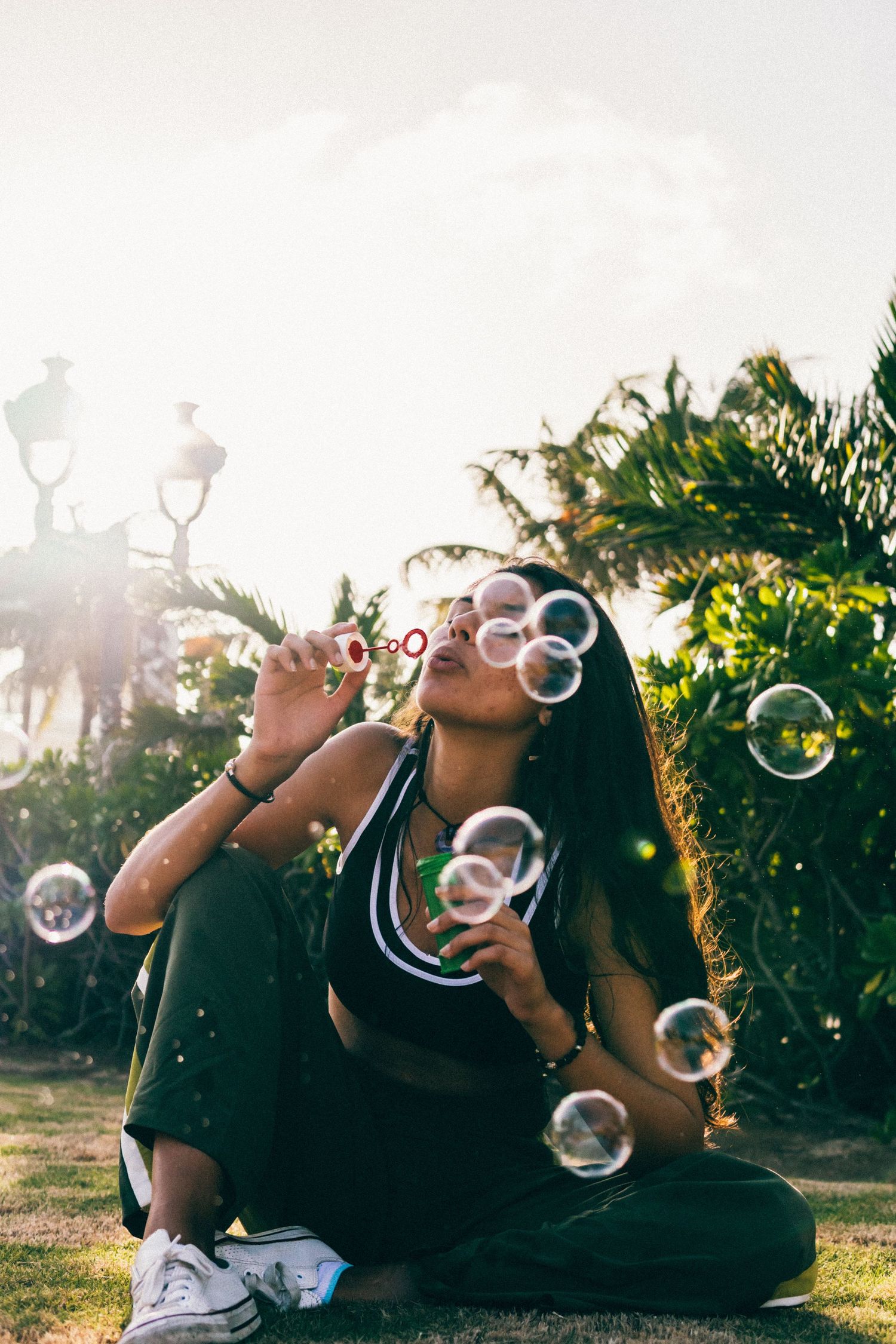 Young woman sitting on grass blowing soap bubbles in a sunlit garden with tropical plants.