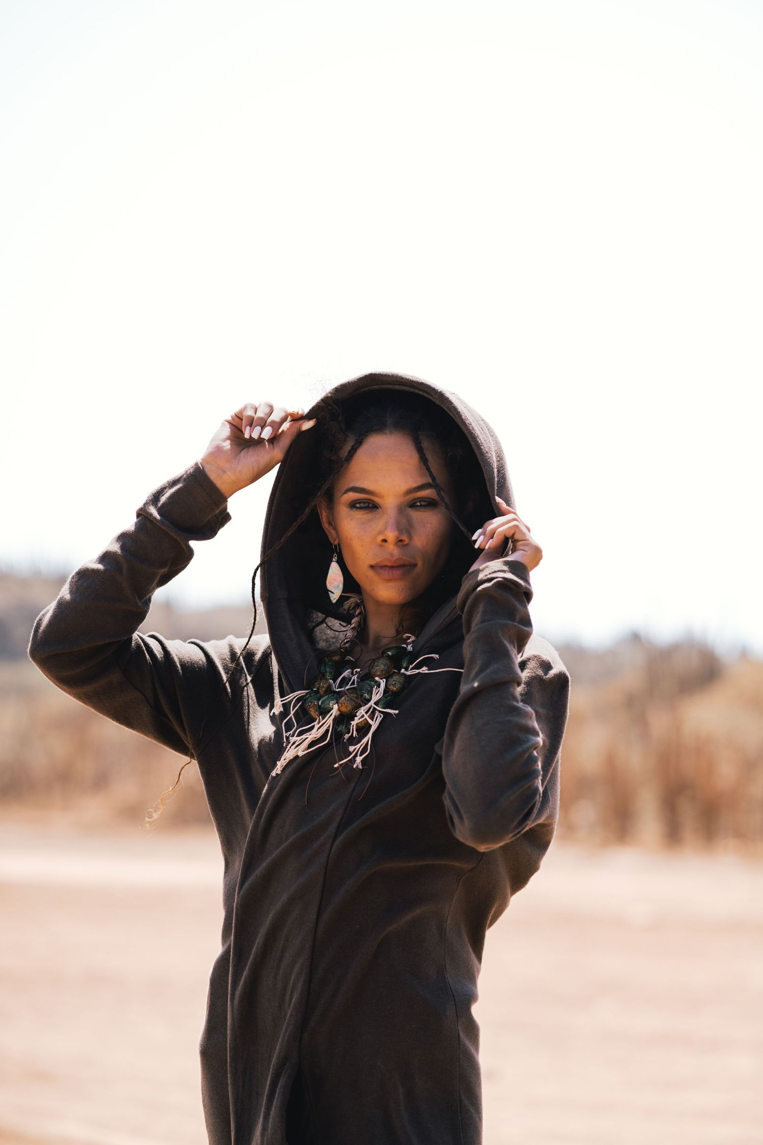 Woman in dark hoodie posing outdoors in arid landscape with hands on hood.