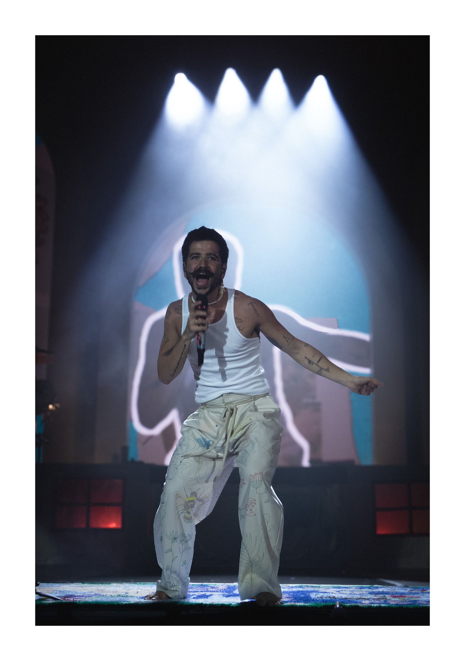 Energetic male performer in white tank and painted pants singing on a blue-lit concert stage.