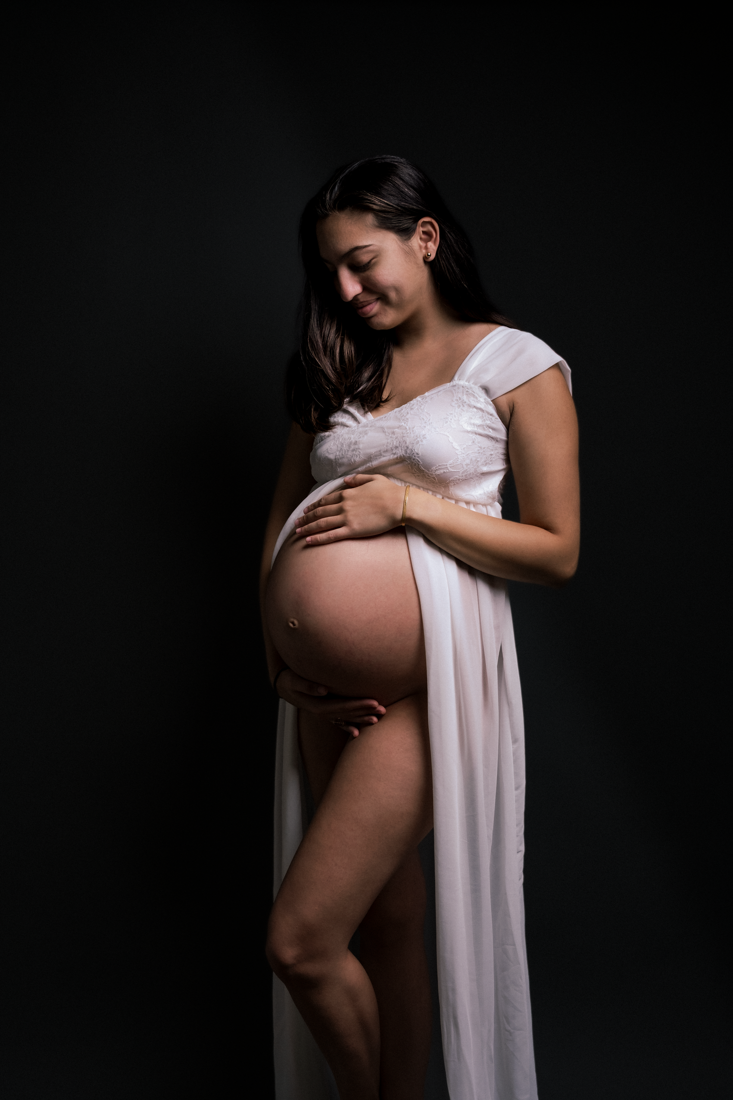 Pregnant woman in white draped fabric cradling her bare belly against a dark studio background.