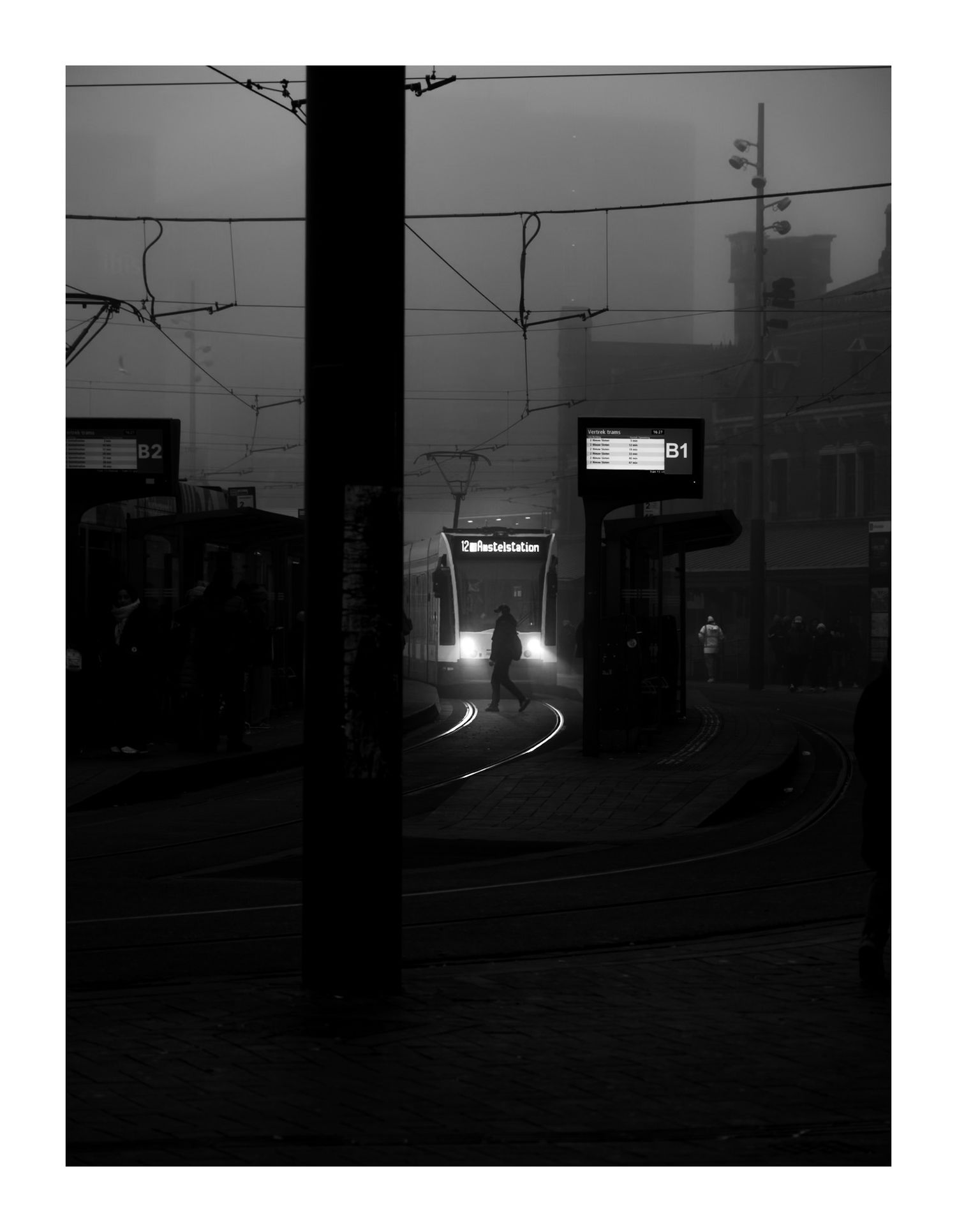 Foggy tram station at night with silhouetted figure crossing tracks, black and white.