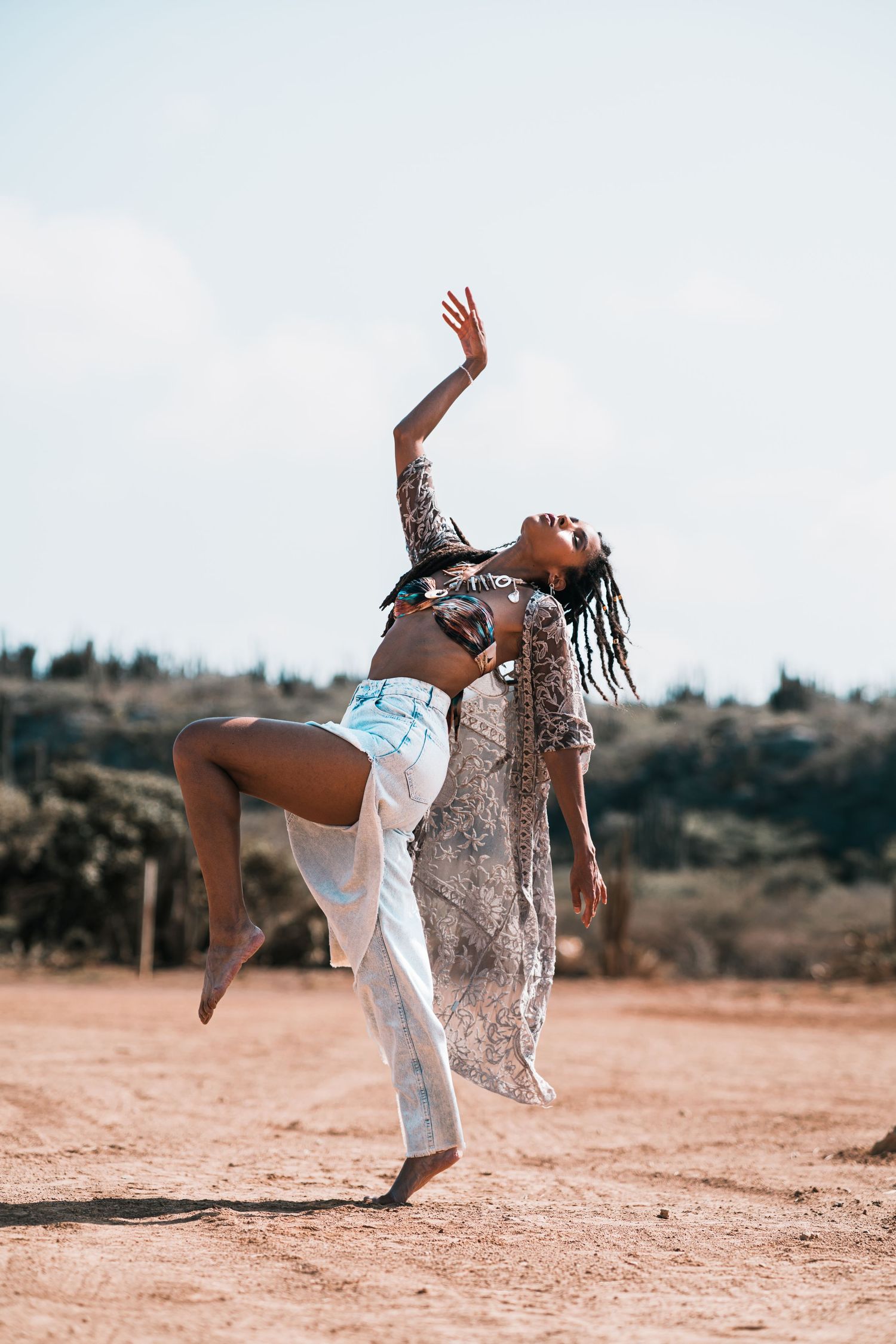 Woman in bohemian attire dancing gracefully on sandy terrain under blue sky.