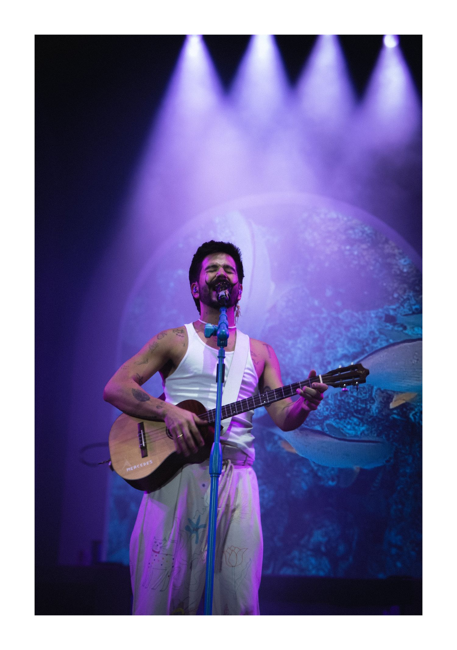 Male musician in white tank top playing acoustic guitar on a purple-lit stage with circular backdrop.
