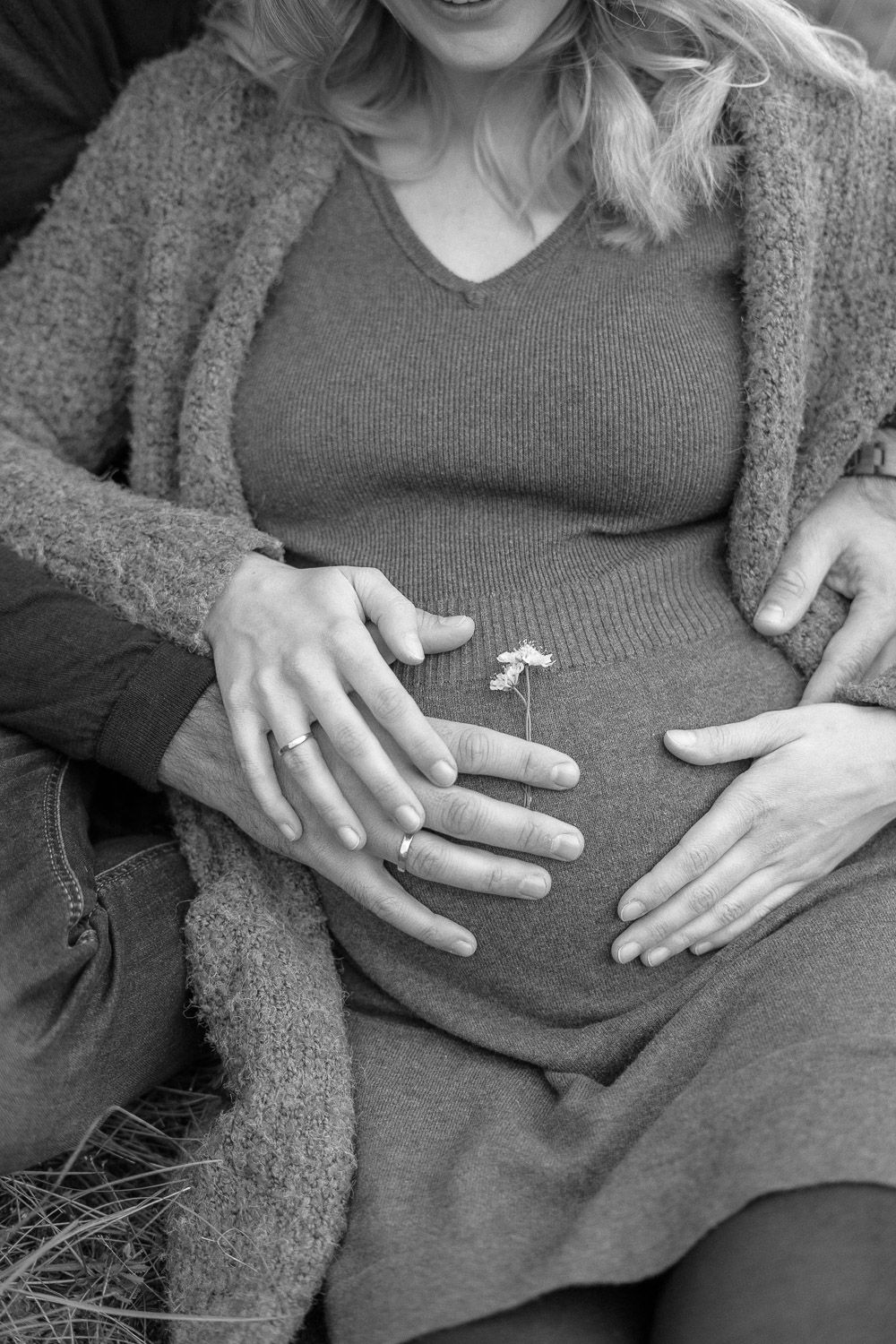 Schwarz-weiß-Nahaufnahme eines Babybauchs, während das Paar auf einer Wiese sitzt.