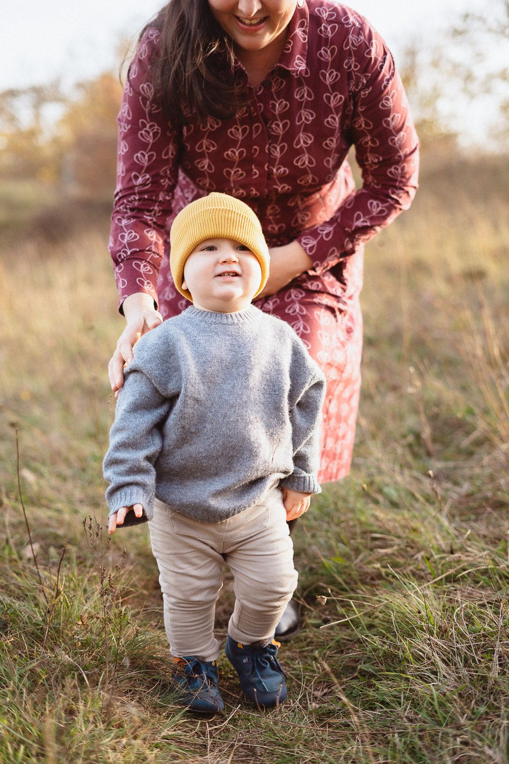 Kleiner Junge mit Mütze, während seine Mutter im Hintergrund liebevoll lächelt