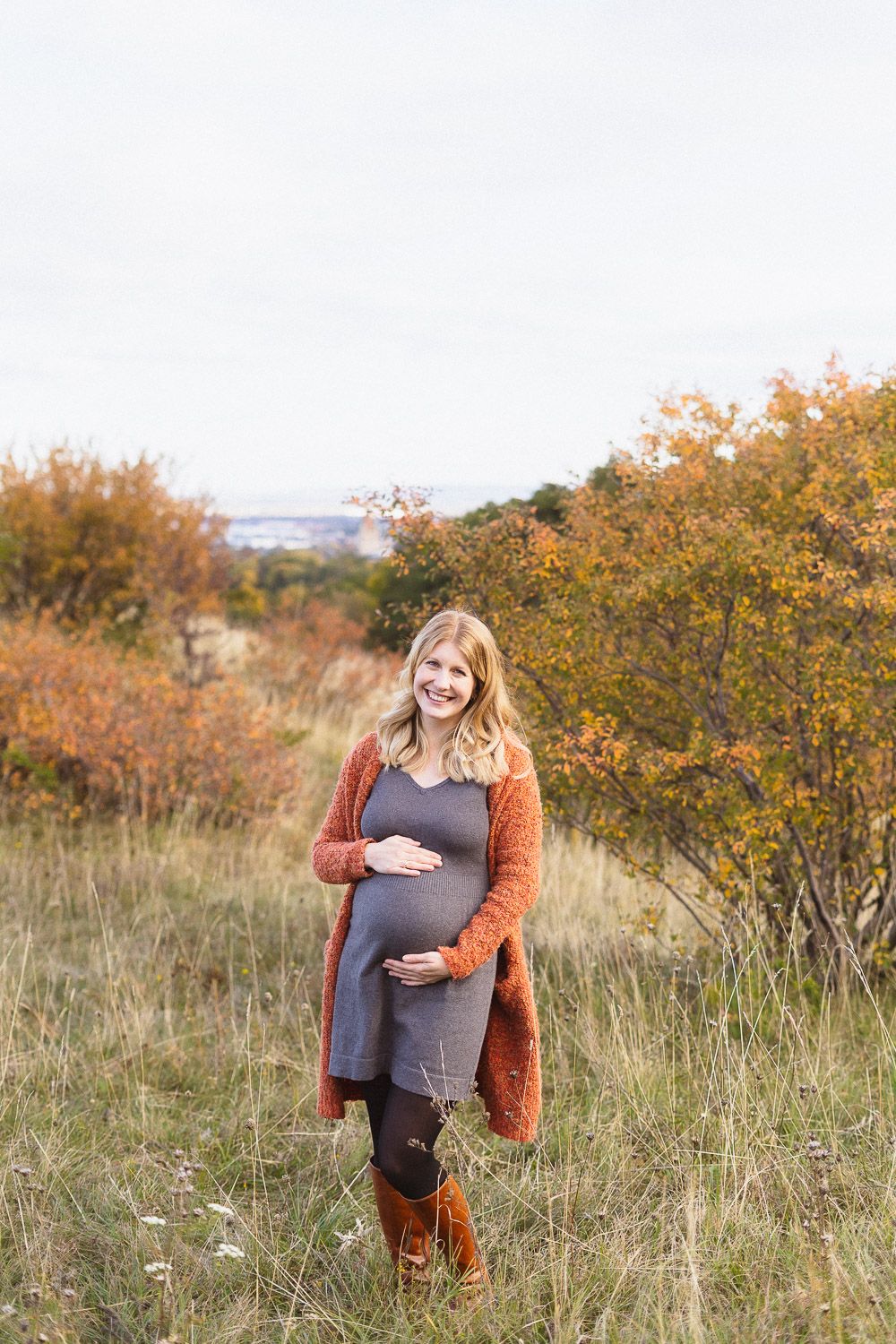 Schwangere Frau beim Babybauchshooting in herbstlicher Natur, hält ihren Babybauch und lächelt in die Kamera.