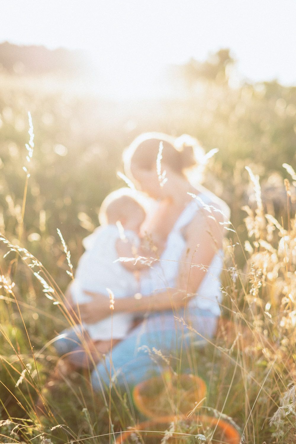 Unscharfes Bild einer Mutter, die ihr Baby stillt, aufgenommen durch Gräser im warmen Gegenlicht