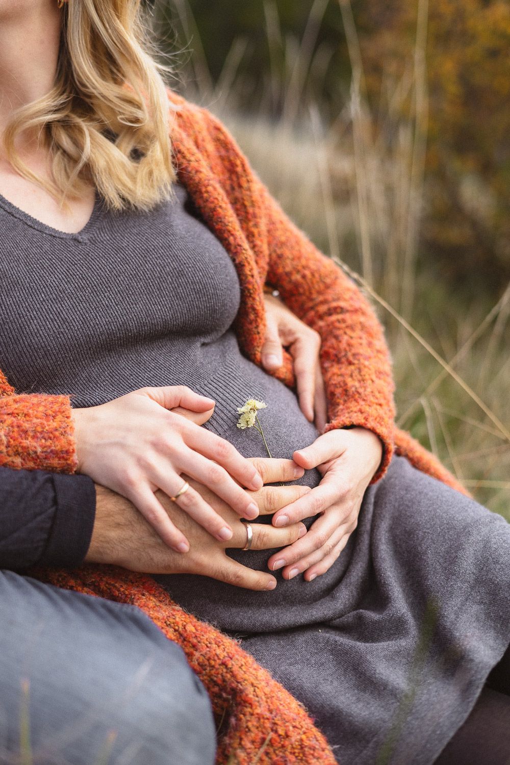 Nahaufnahme eines Babybauchs, auf dem eine Blume liegt, während das Paar auf einer Wiese sitzt.