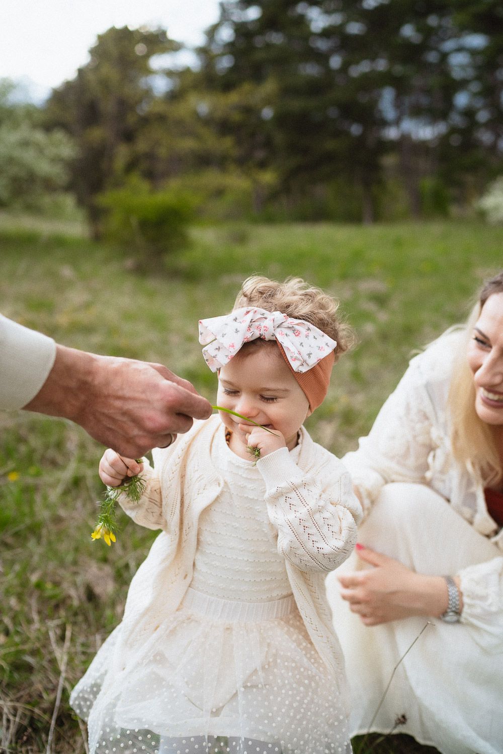 Vater kitzelt seine Tochter mit einer Blume an der Nase, während die Mutter im Hintergrund lächelt.