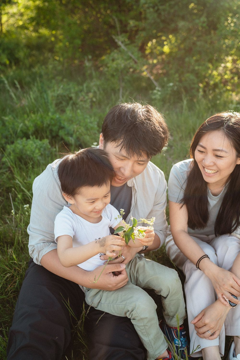 Familienshooting in der Natur – Eltern sitzen mit ihrem Sohn auf der Wiese und betrachten gemeinsam eine Blume in seinen Händen.