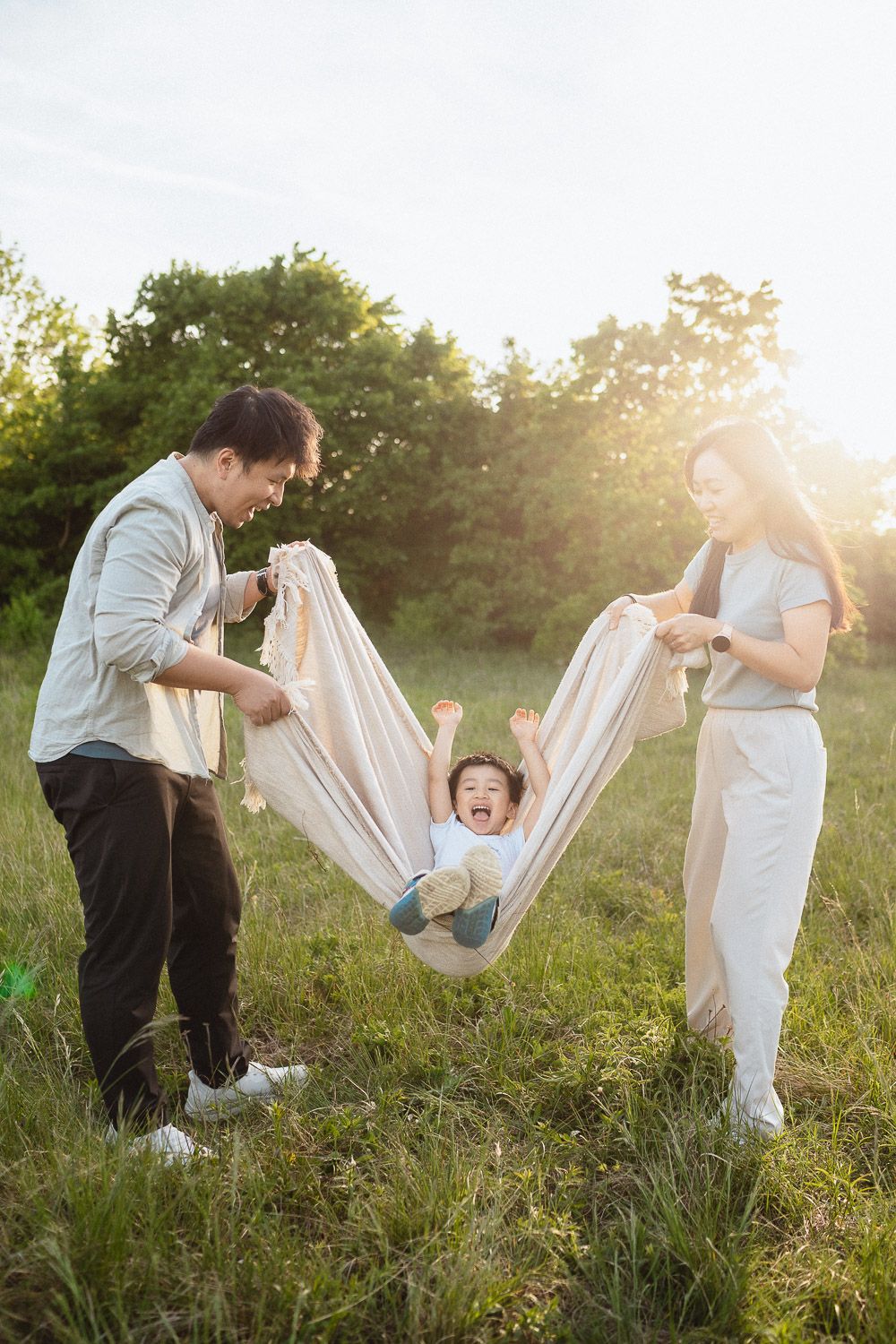 Familienshooting im Abendlicht – Eltern schaukeln ihren lachenden Sohn in einer Decke.