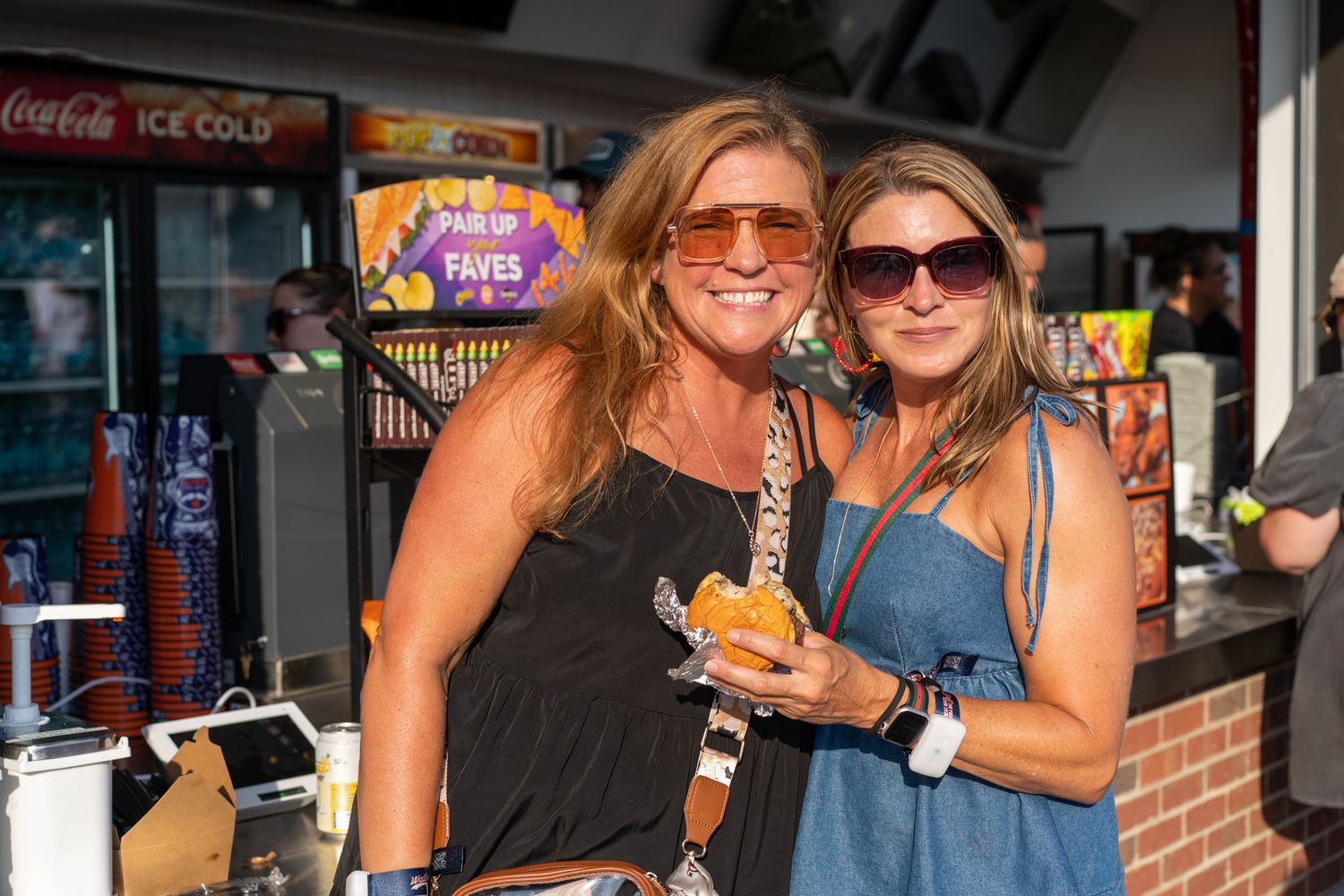 Two women smiling and posing together at an outdoor food stand, one holding a snack, on a sunny day.