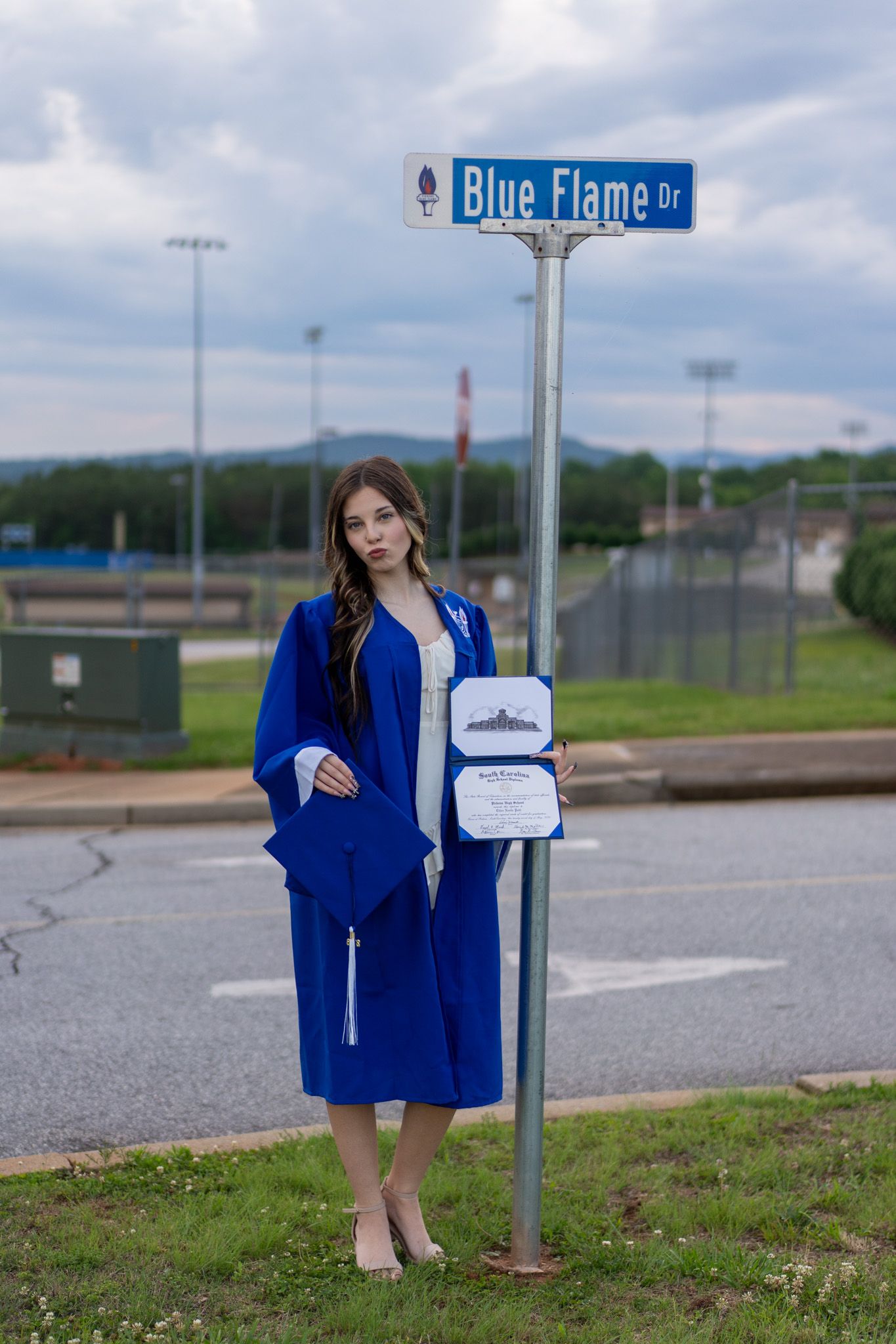 Young woman in blue graduation cap and gown holding diploma near Blue Flame Dr street sign.