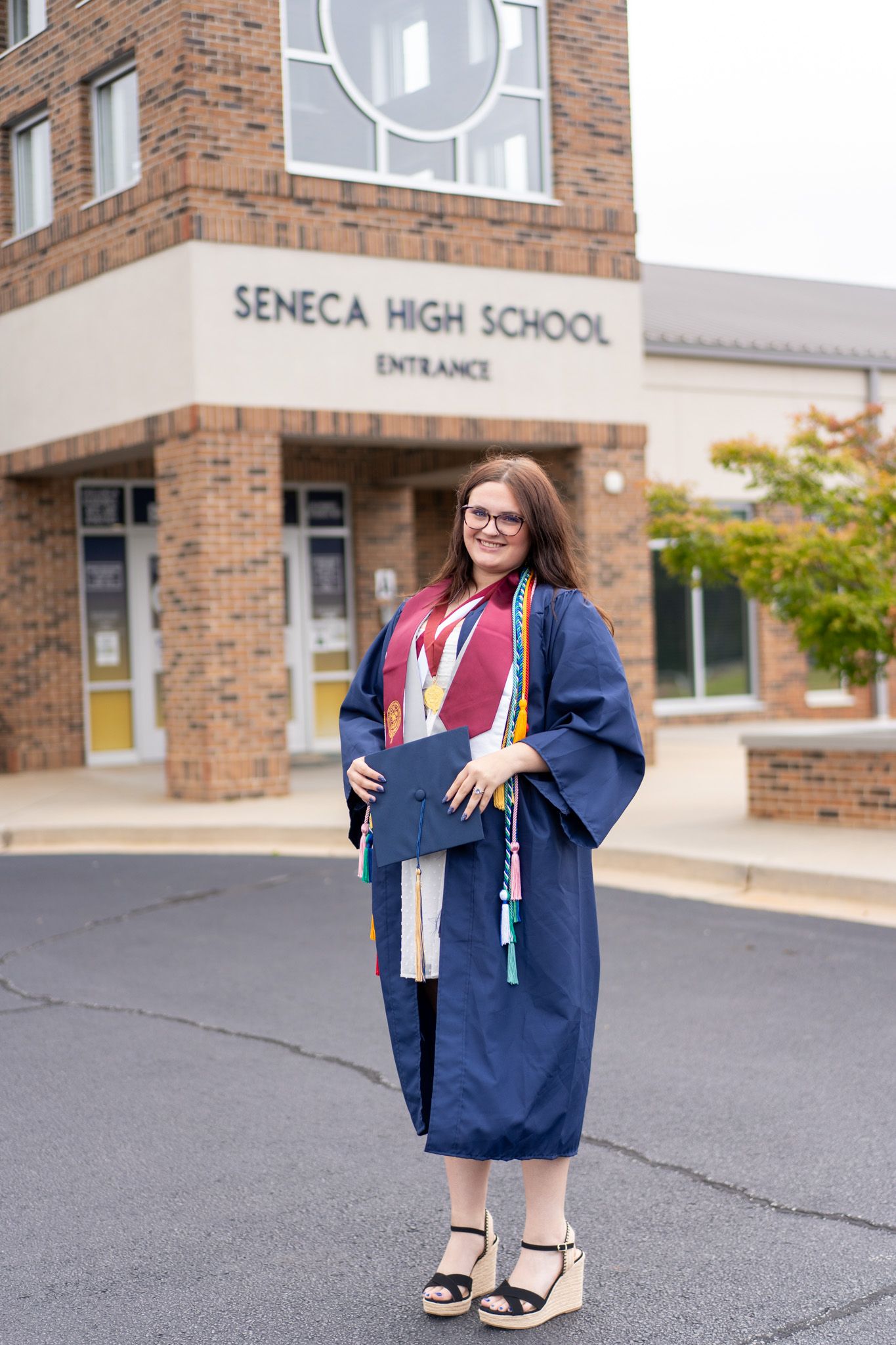A smiling graduate in navy cap and gown poses outside Seneca High School entrance holding her diploma.