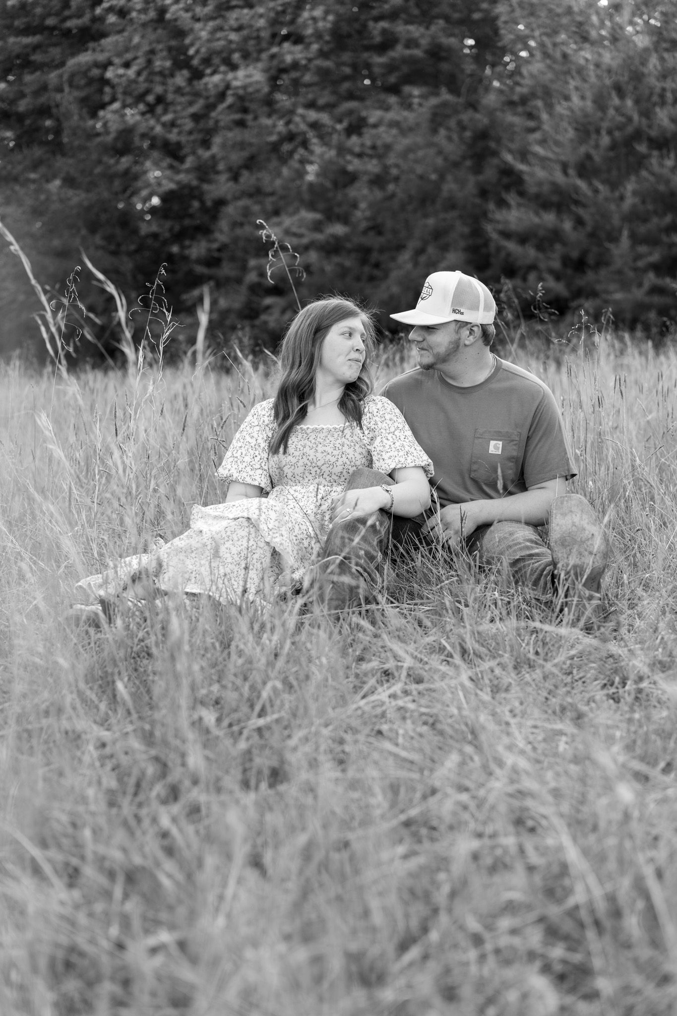 Black and white photo of a couple sitting together in tall grass field, gazing at each other during an outdoor engagement session.
