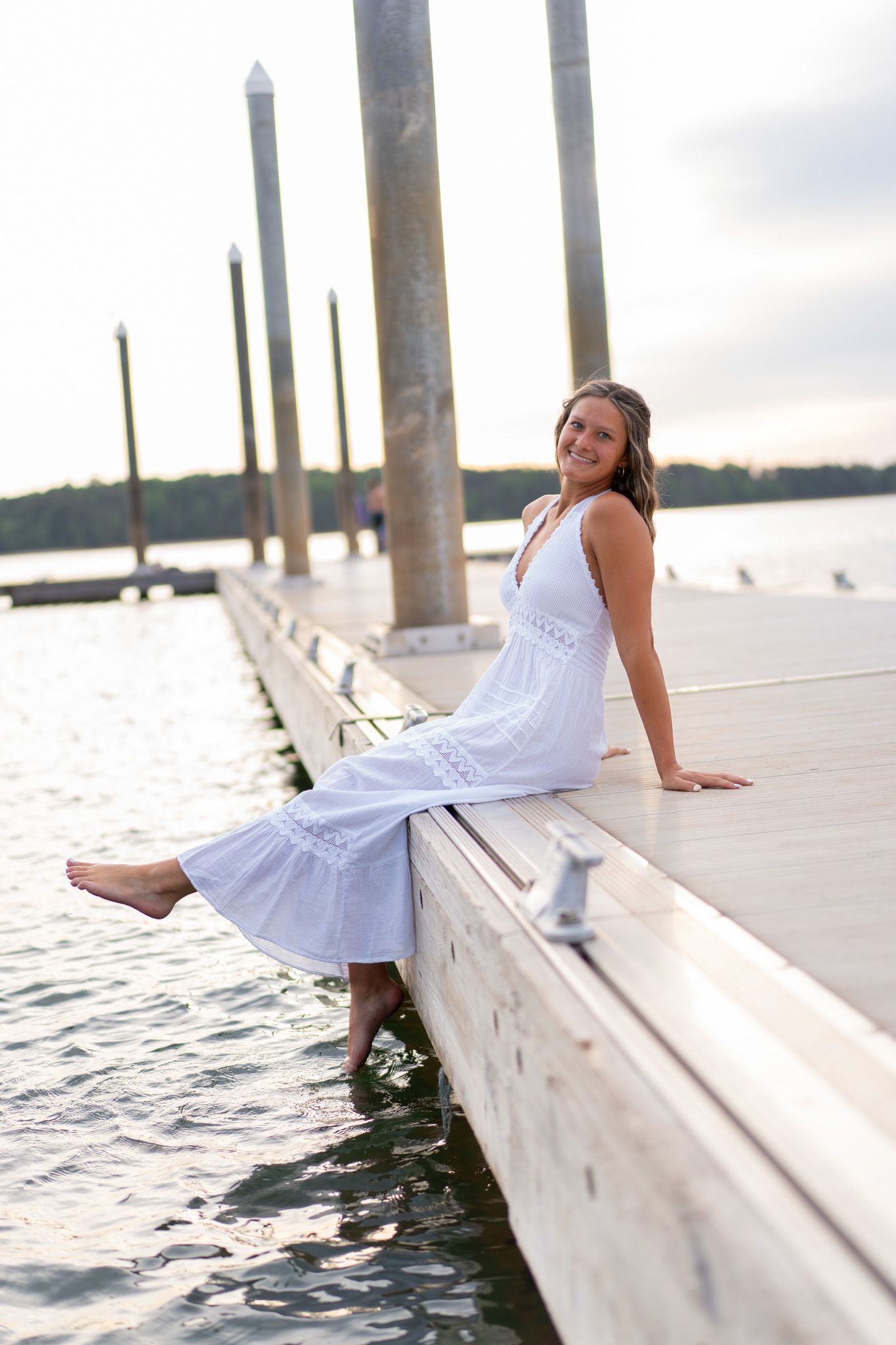 Smiling young woman in white dress sitting on wooden dock by calm lake at sunset.