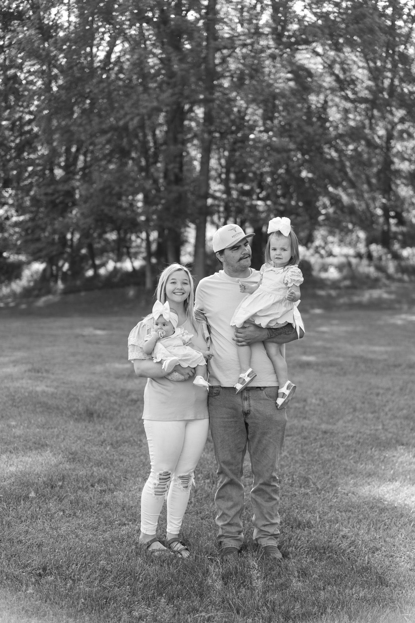 Black and white family portrait of parents holding newborn and toddler in a park setting.