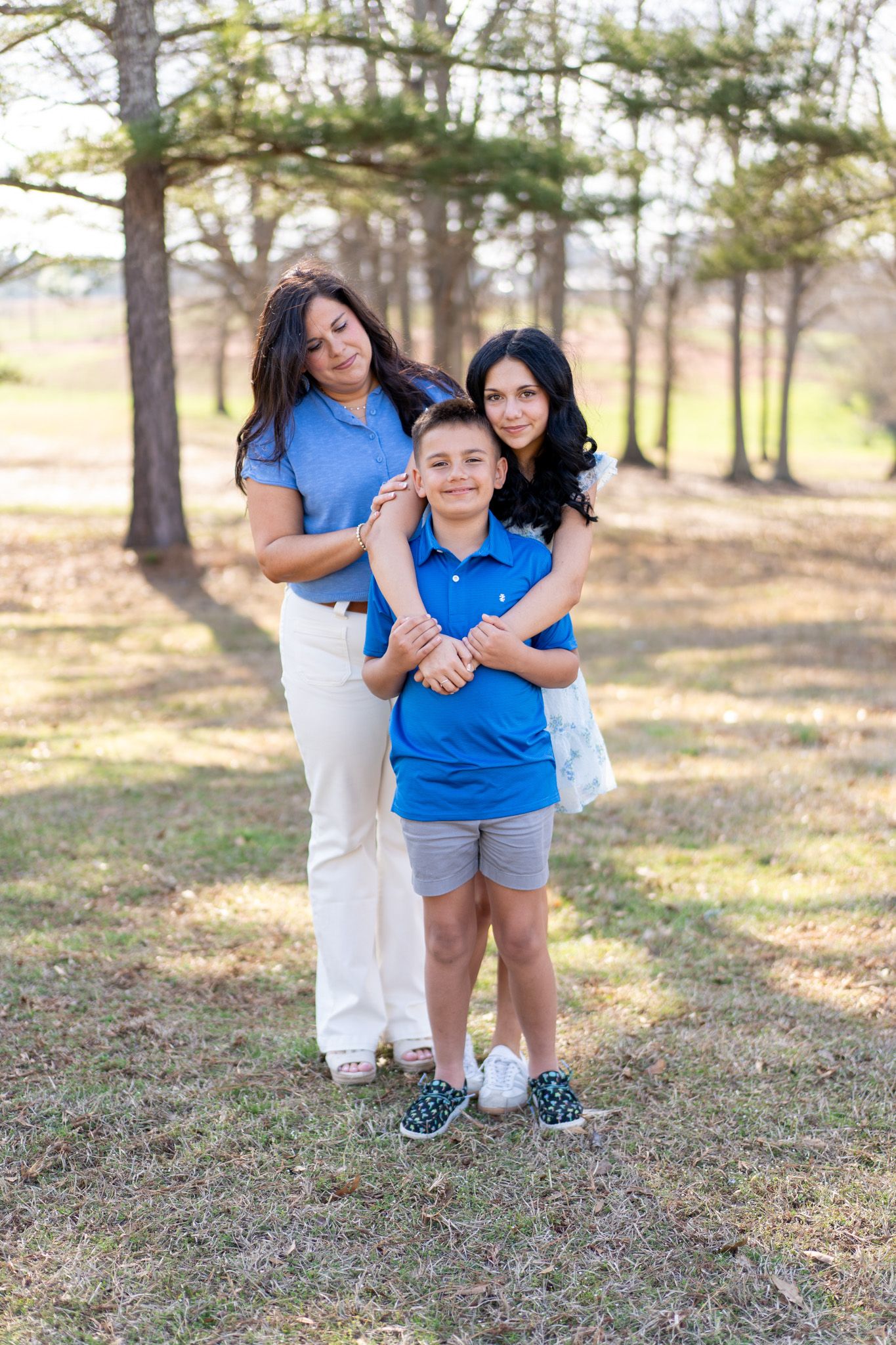 Two women and a boy in blue polo posing together in a tree-lined outdoor setting.