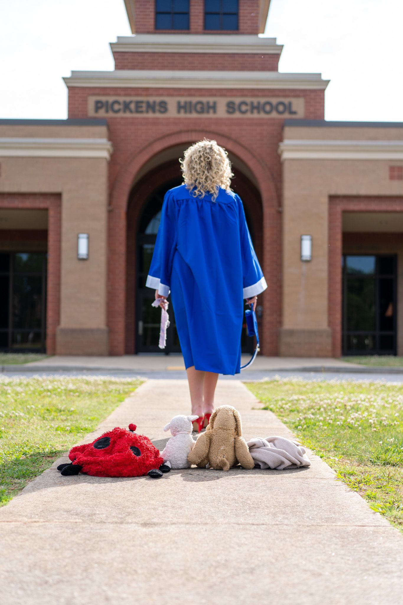 A graduate in blue cap and gown stands before Pickens High School with childhood stuffed animals on the walkway.