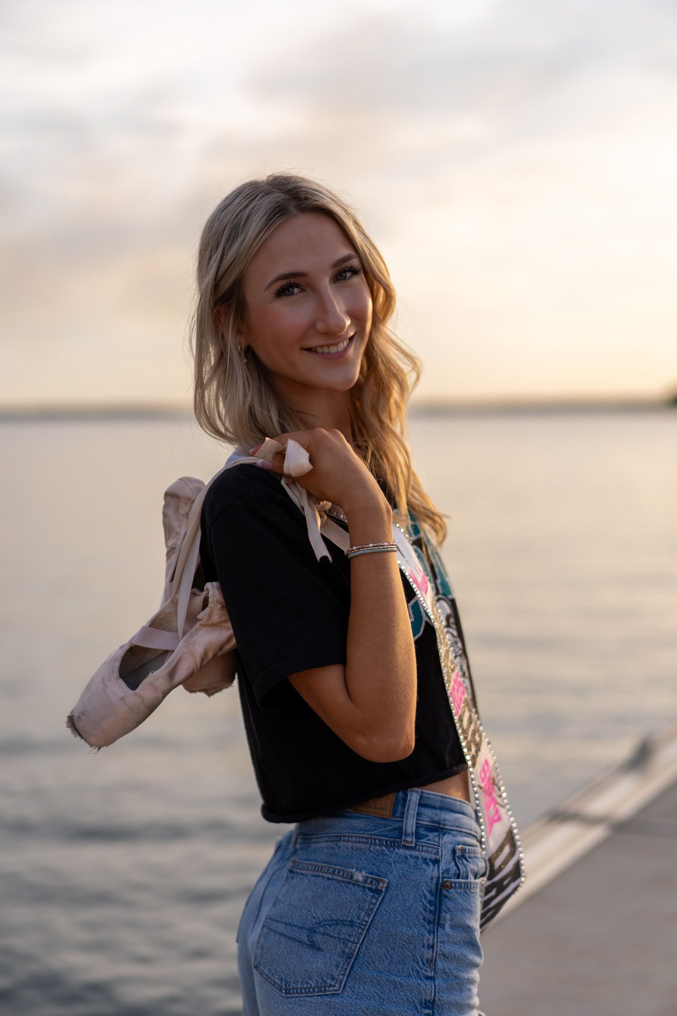 Smiling blonde woman in black crop top and jeans holding ballet shoes by waterfront at sunset.