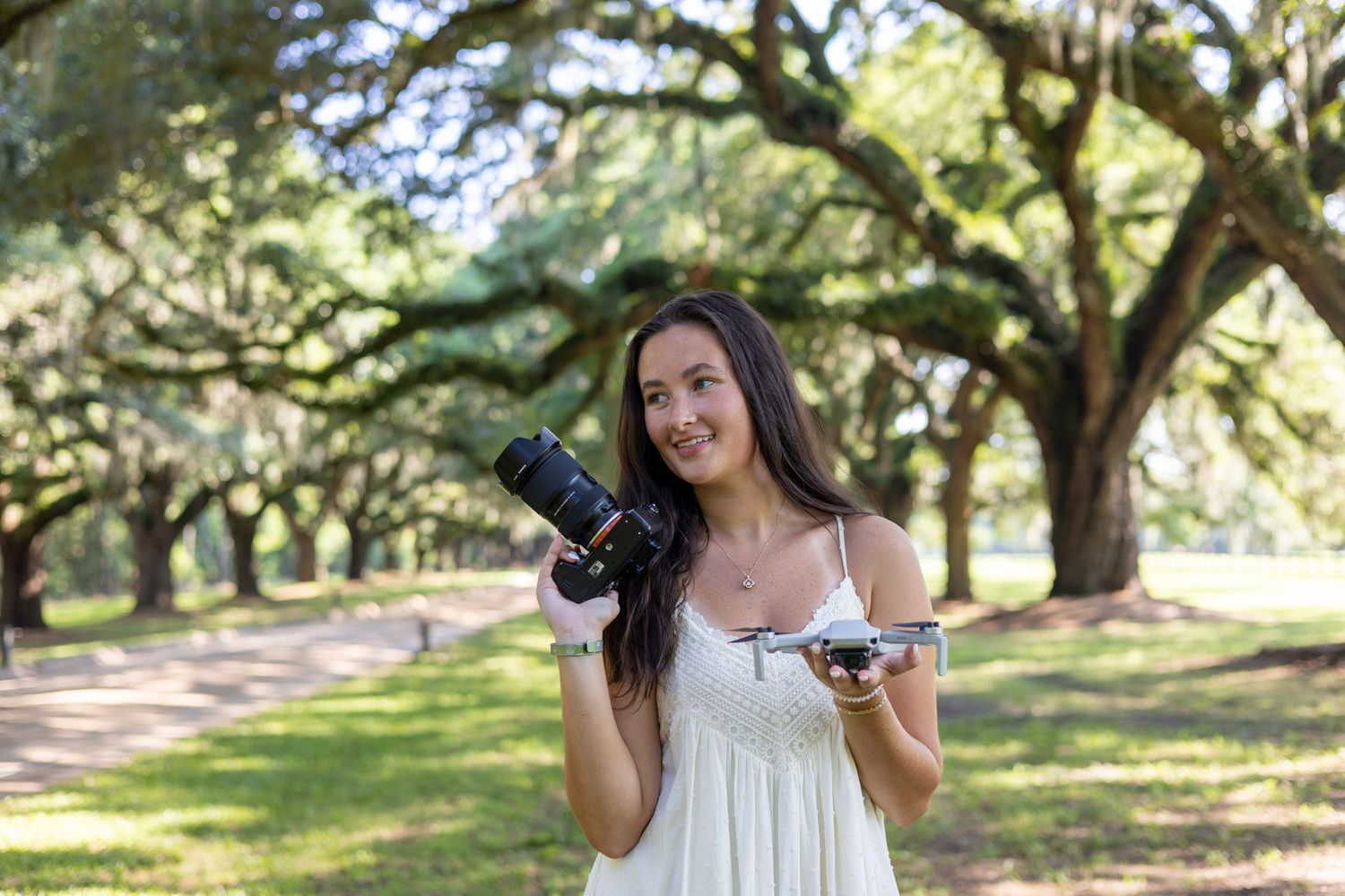 Female wedding photographer smiles holding DSLR camera and drone under oak tree canopy.