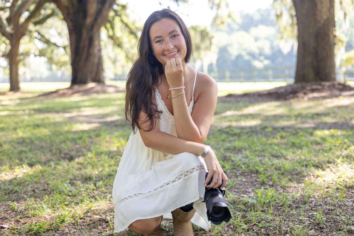 Smiling woman in white dress crouching with camera among large oak trees in a sunlit outdoor setting.