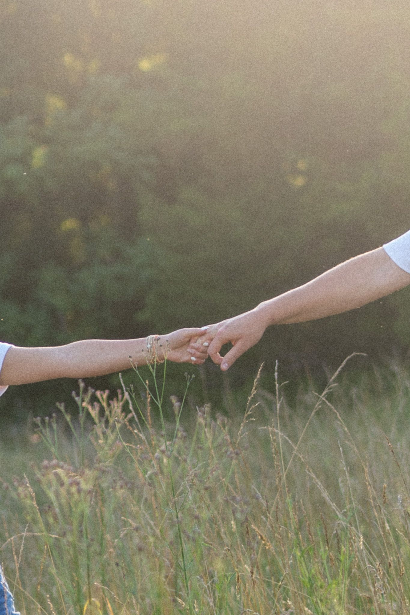 Close-up of couple gently reaching for each other's hands in a sunlit grassy field.