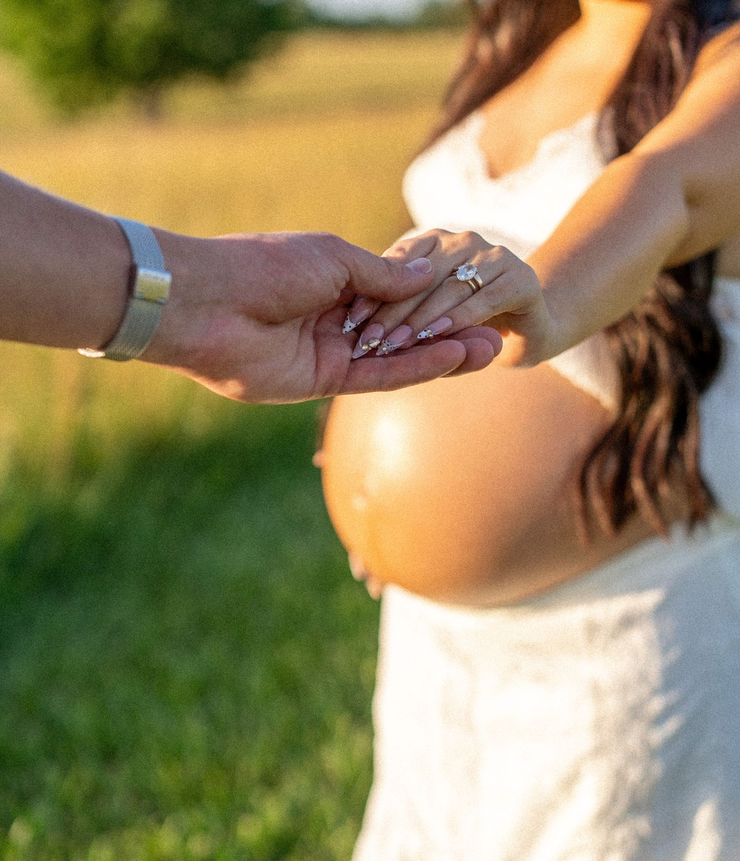 Couple holding hands outdoors highlighting diamond engagement ring on pregnant woman's finger.