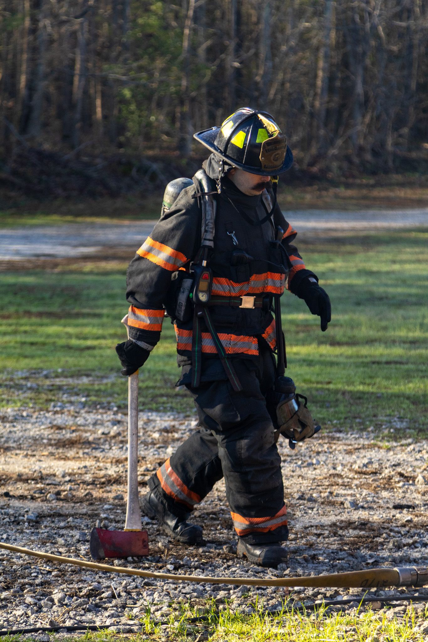 Firefighter in black turnout gear carrying an axe while walking across gravel at an outdoor scene.
