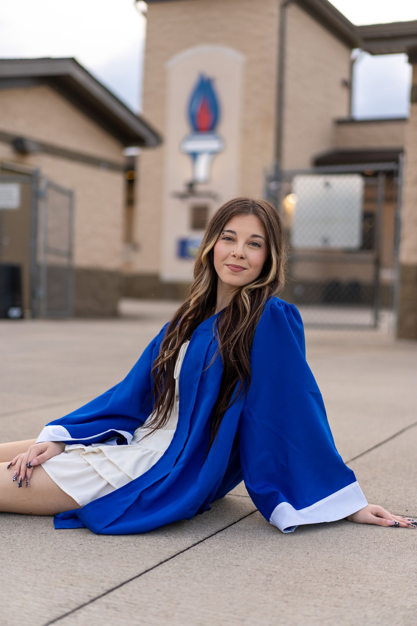 High school senior girl in blue graduation gown sits on ground outside school building for portrait.
