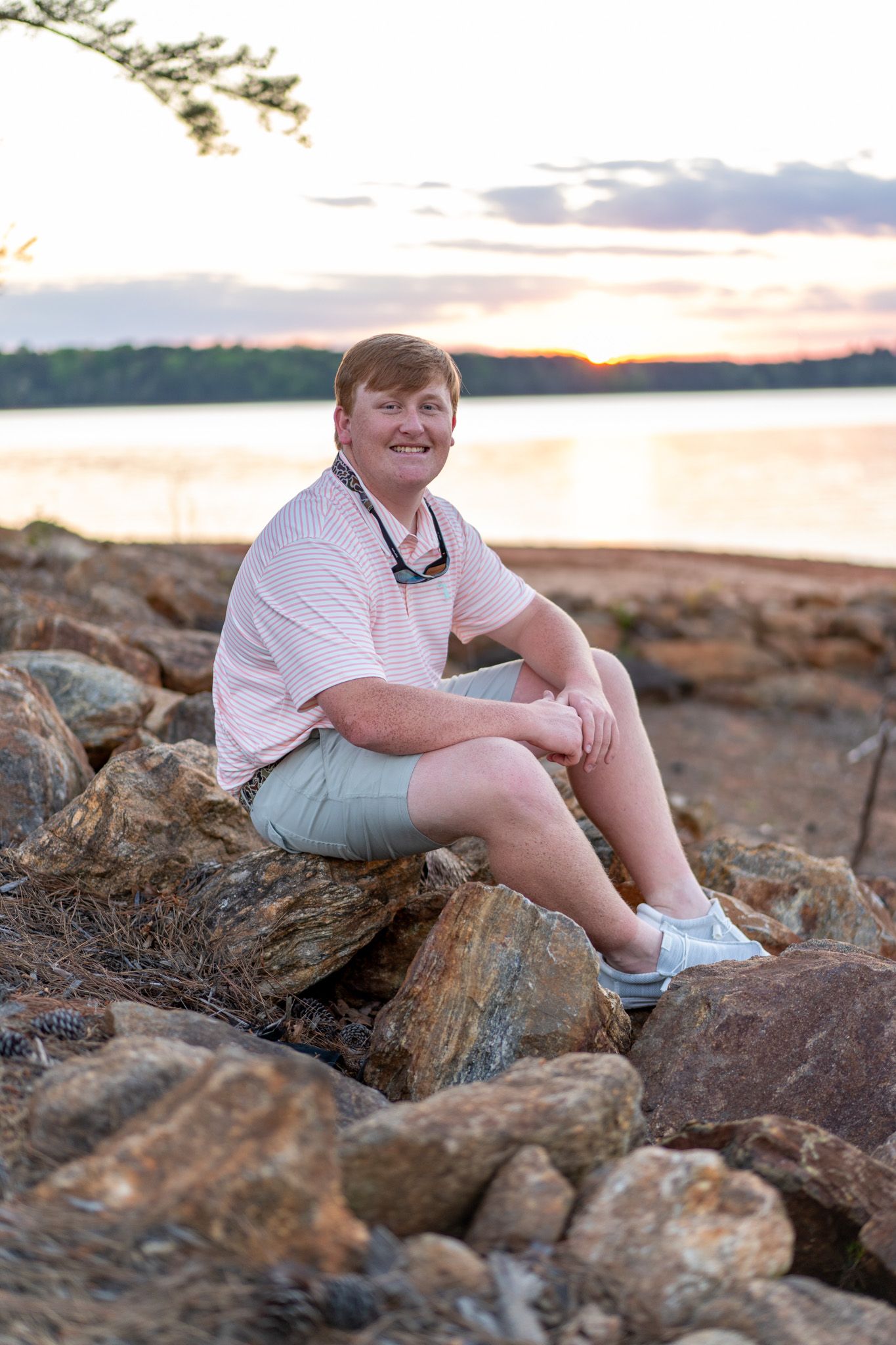 Smiling person sitting on rocky shoreline at sunset, wearing pink shirt and gray shorts, with calm lake in background.
