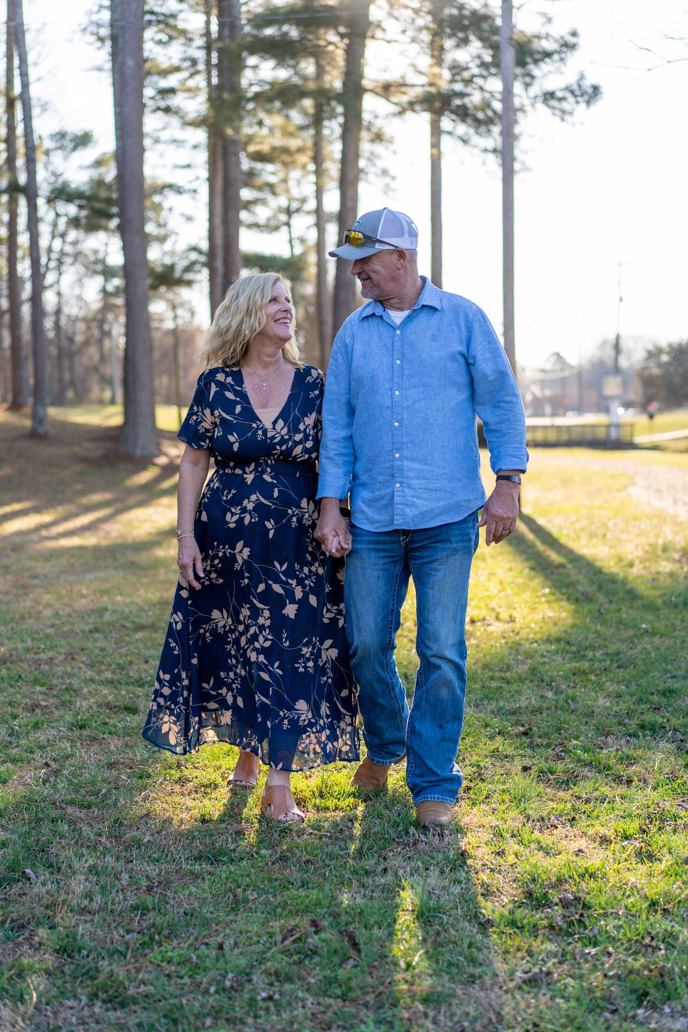 Happy couple holding hands and smiling while walking through a sunny park with tall pines.