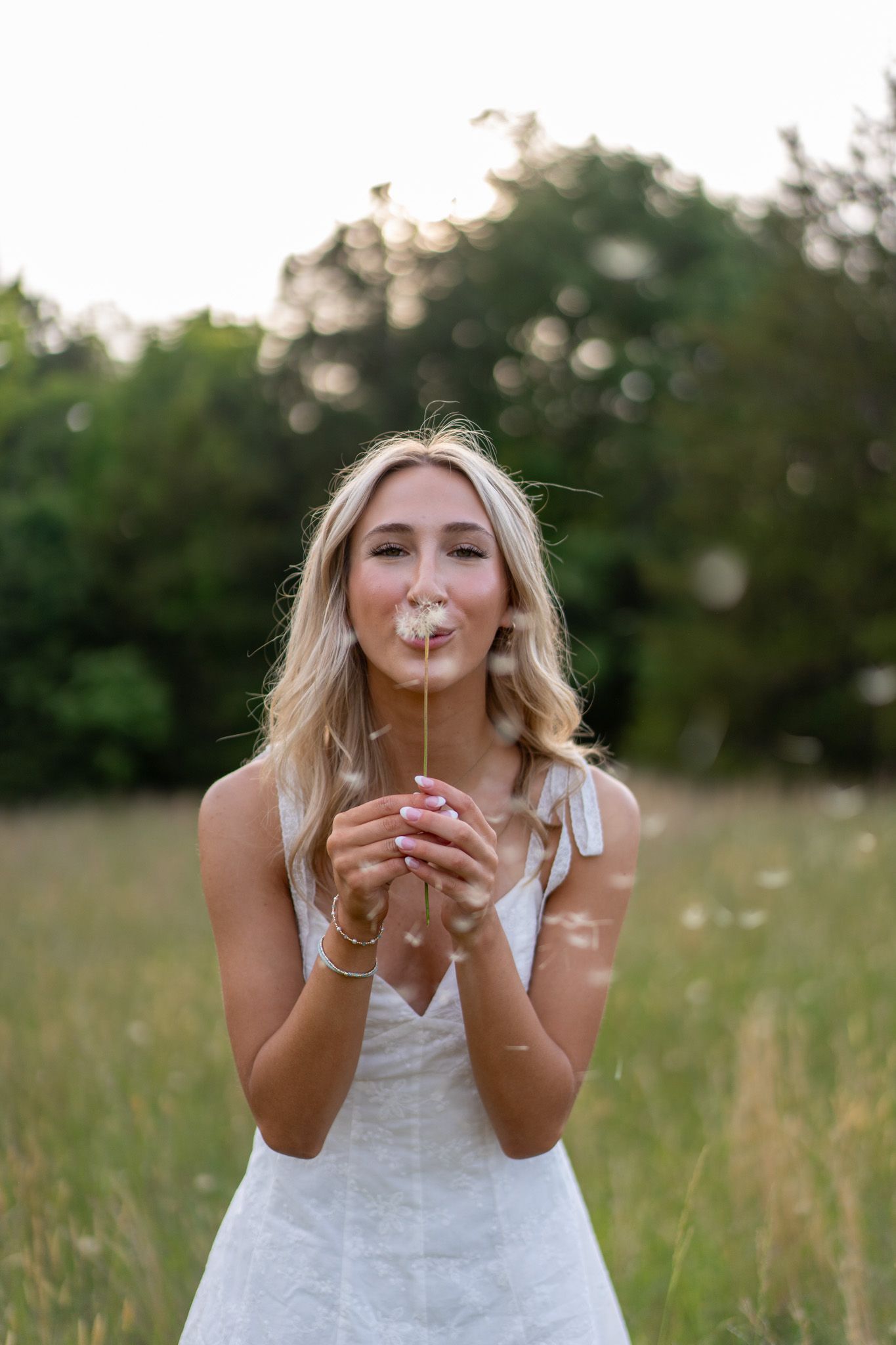 Blonde woman in white dress blowing a dandelion in a sunny green meadow.