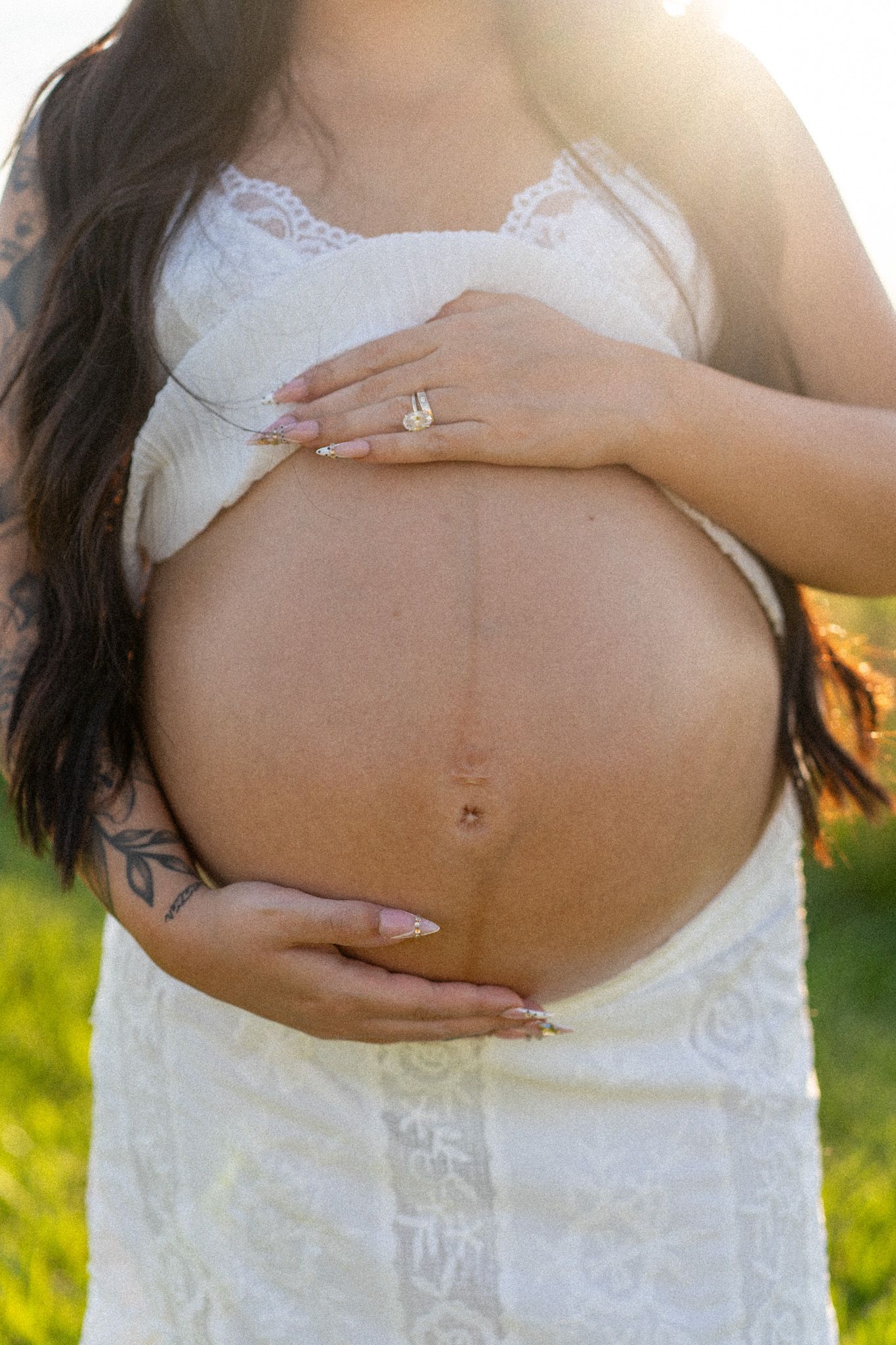 Pregnant woman in white lace dress cradling baby bump outdoors in golden sunlight, showing engagement ring and tattoos.
