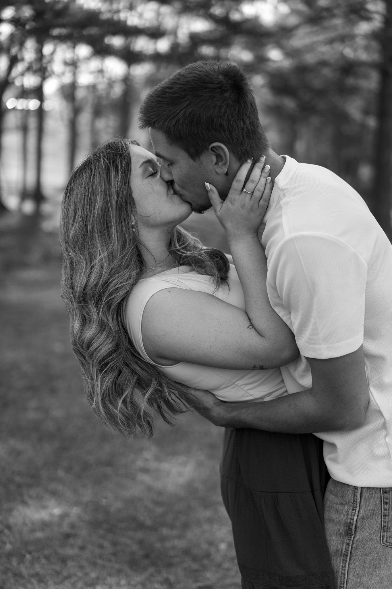 Couple sharing a romantic kiss in black and white photo among tall pine trees.