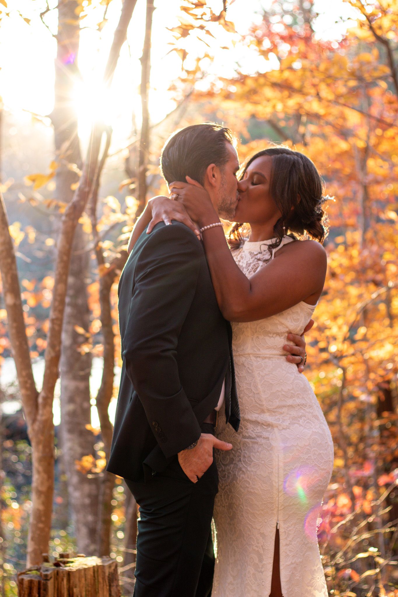 Newlywed couple kissing in golden autumn forest with sunlight streaming through colorful leaves.