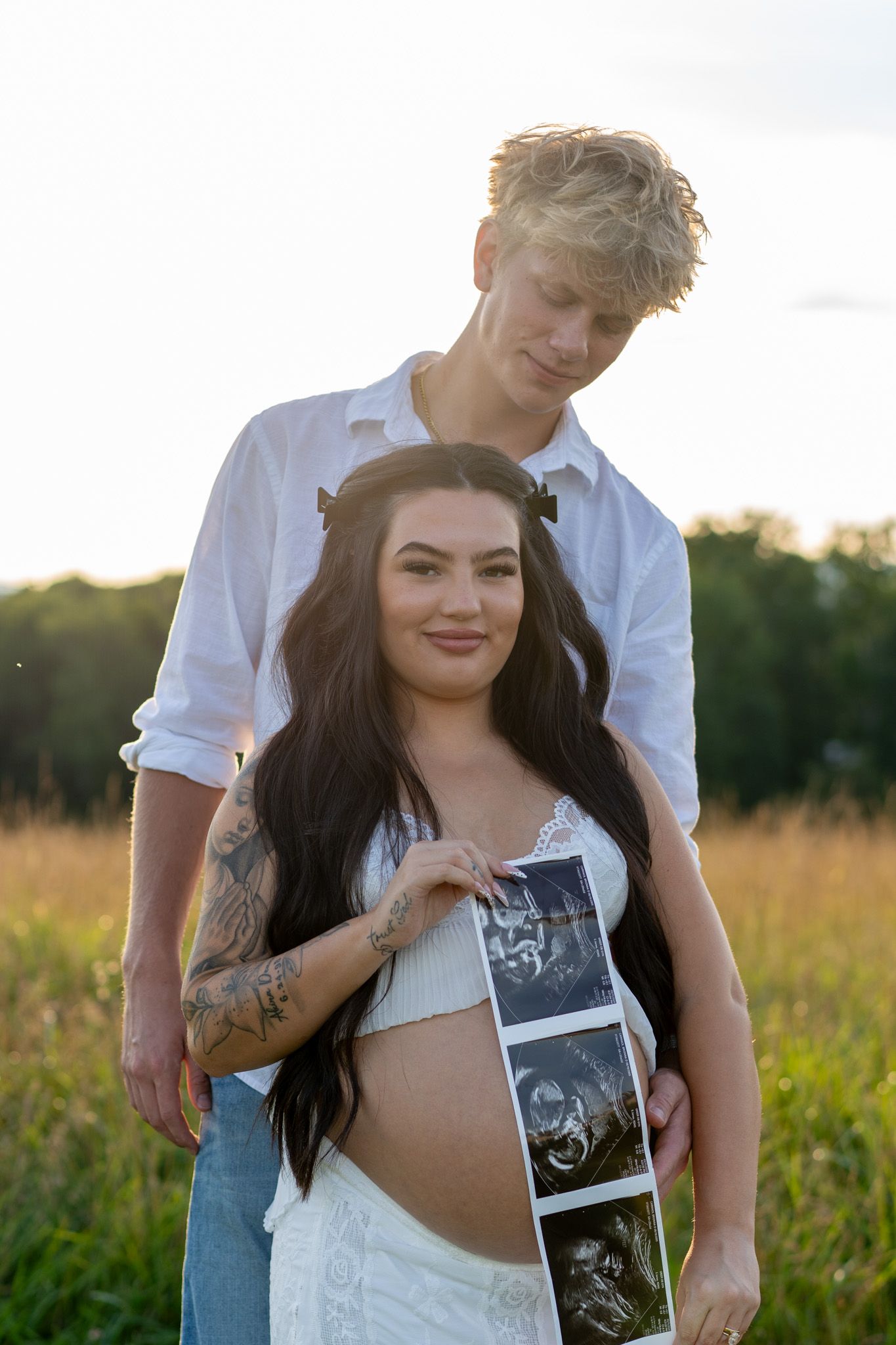 Smiling pregnant woman in white lace dress holding ultrasound photos with blonde partner in sunny field.