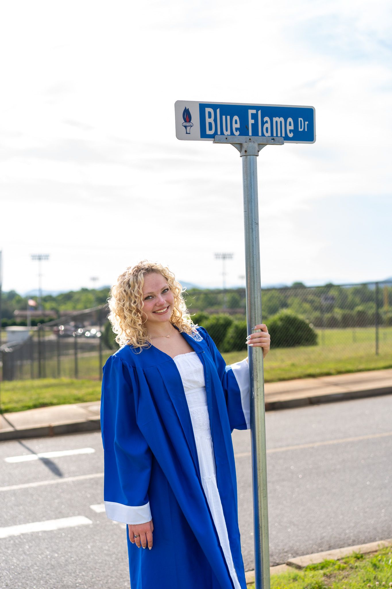 A smiling graduate in a blue cap and gown poses beside a Blue Flame Dr street sign outdoors.