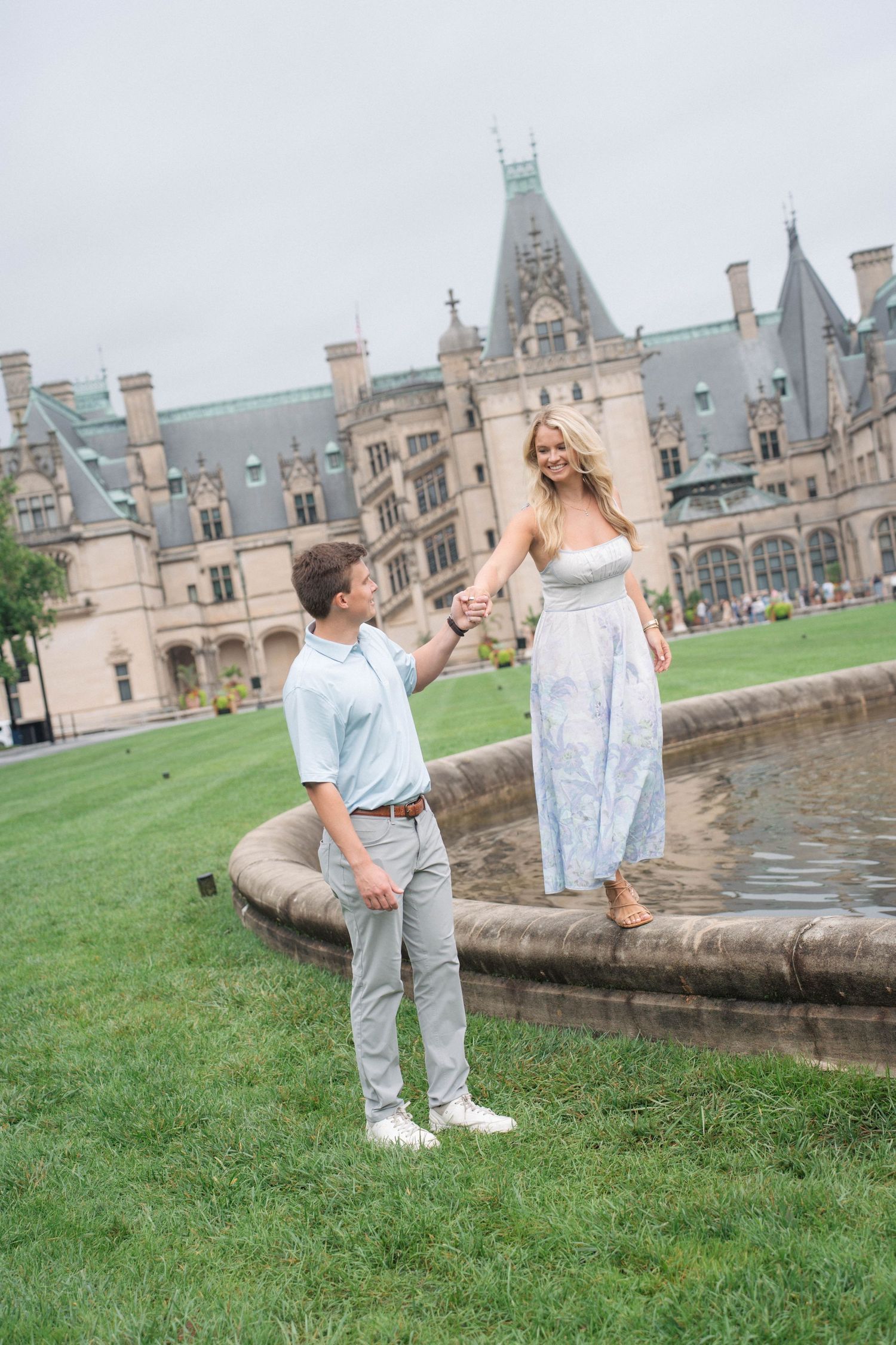 Woman standing on fountain edge holding hands with man on green lawn before historic Biltmore Estate.