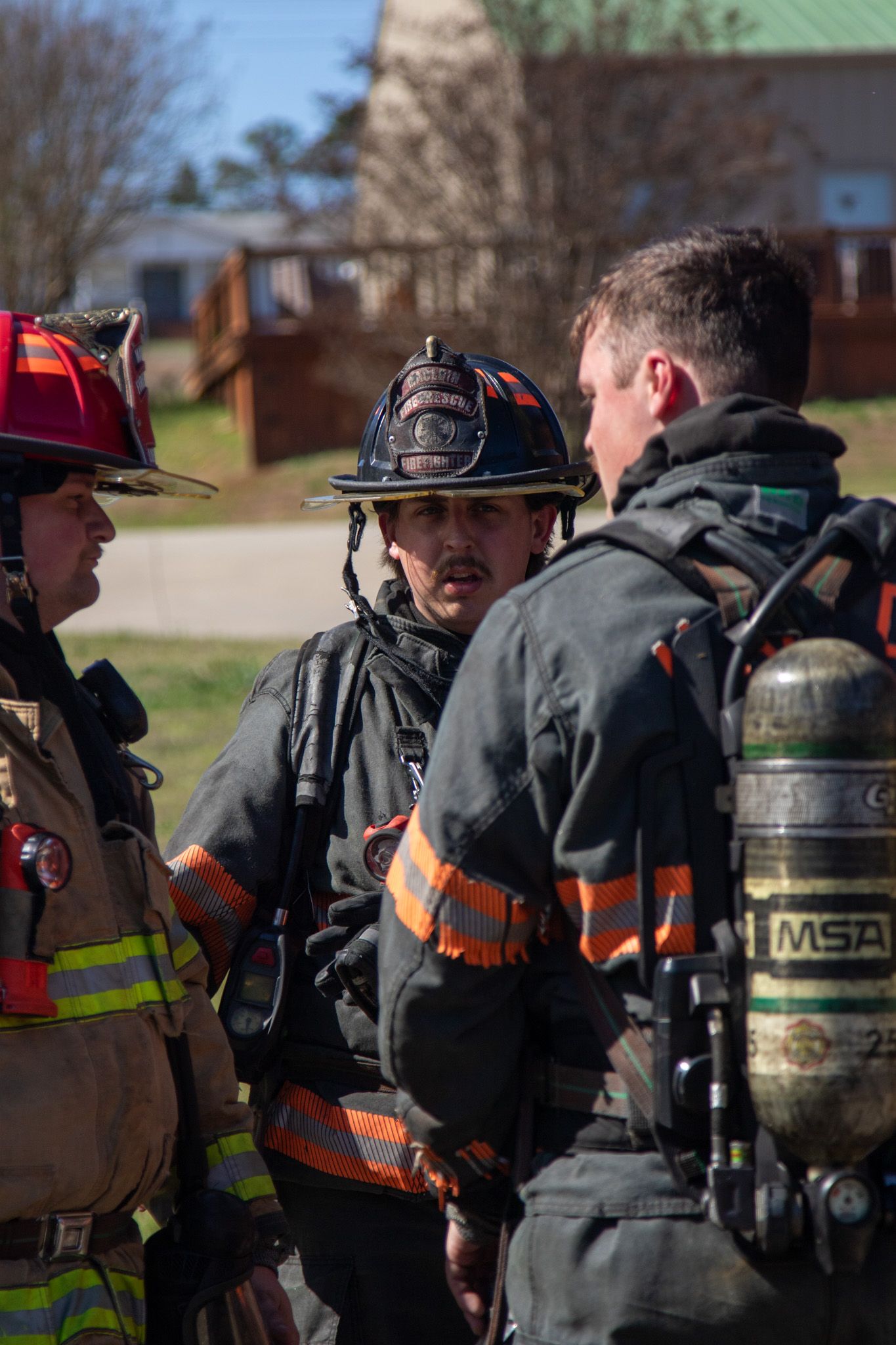 Three firefighters in turnout gear and helmets discuss operations outdoors near residential buildings.