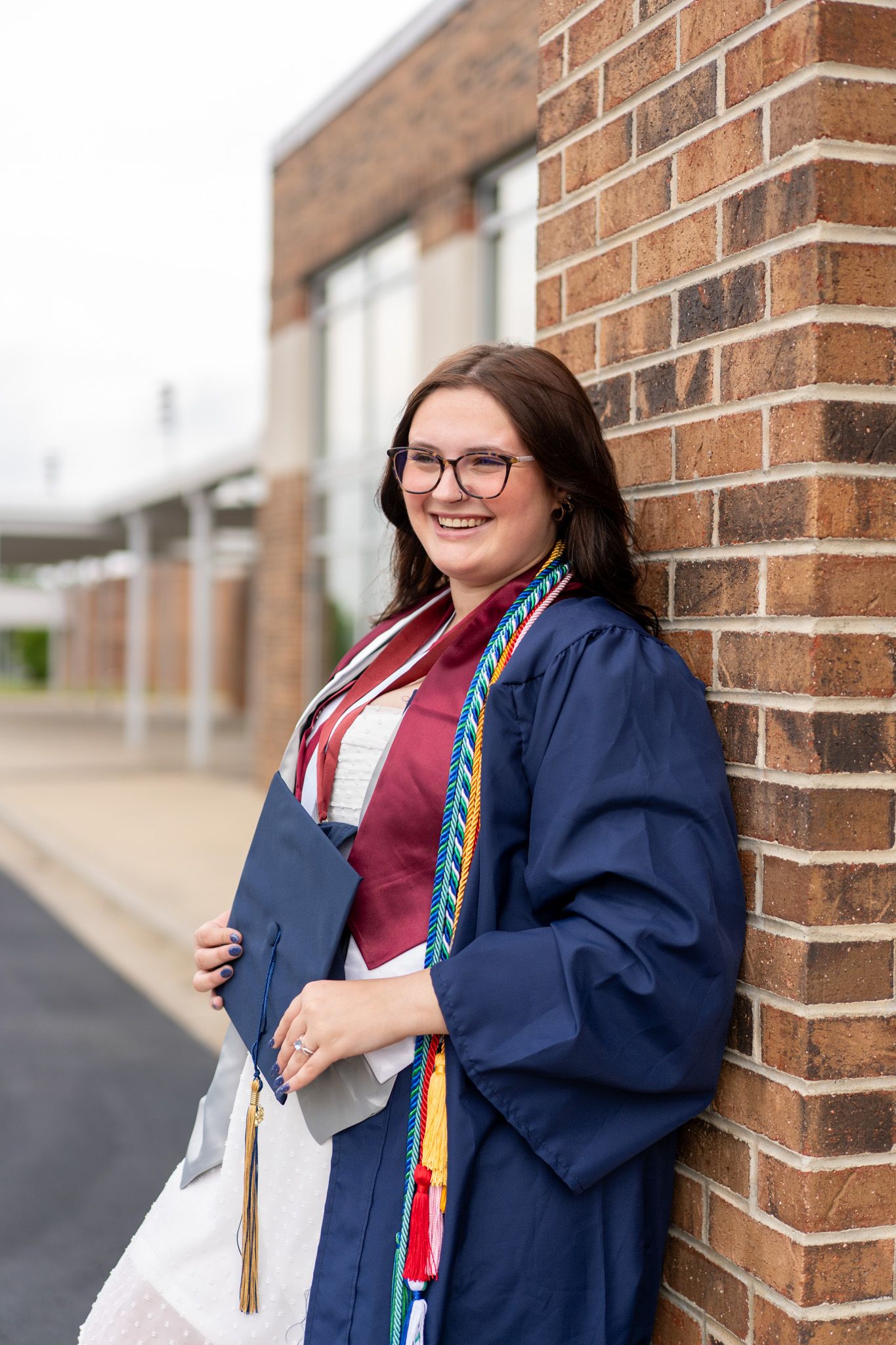Smiling female graduate in navy blue cap and gown with honor cords leaning against brick wall outside school.