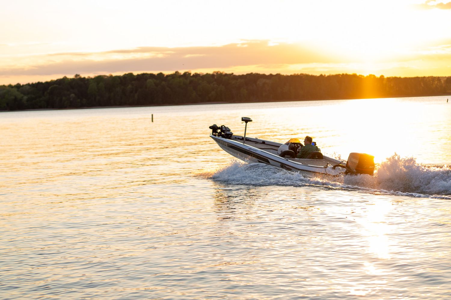 A speedboat races across a calm lake at golden sunset, creating white wake waves with a tree-lined horizon in the background.