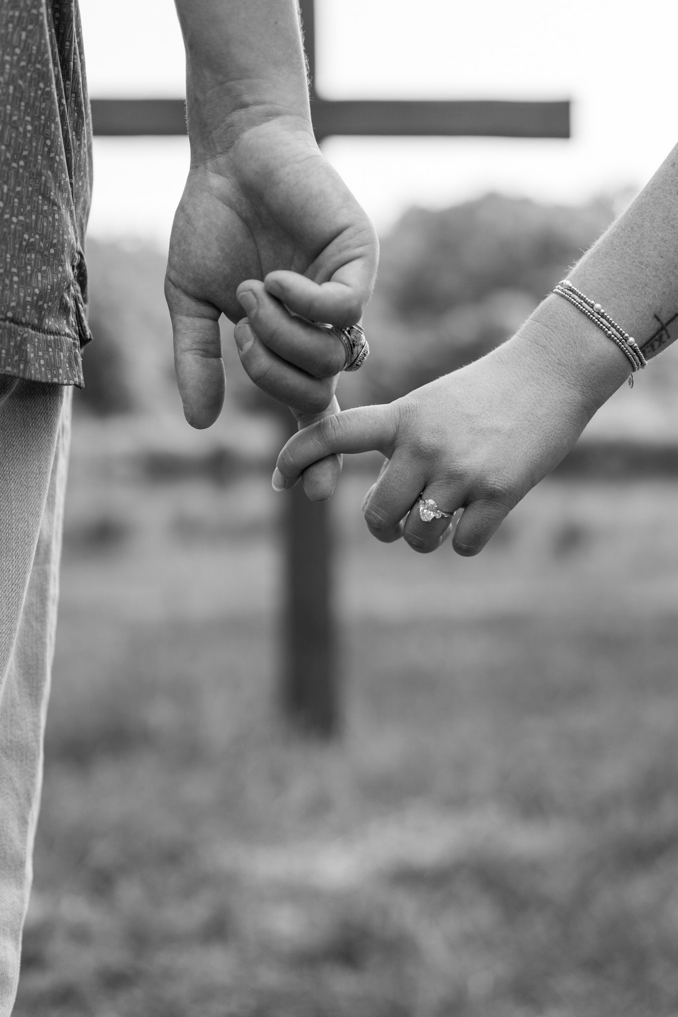Black and white close-up of couple holding hands with a wooden cross visible in the background.
