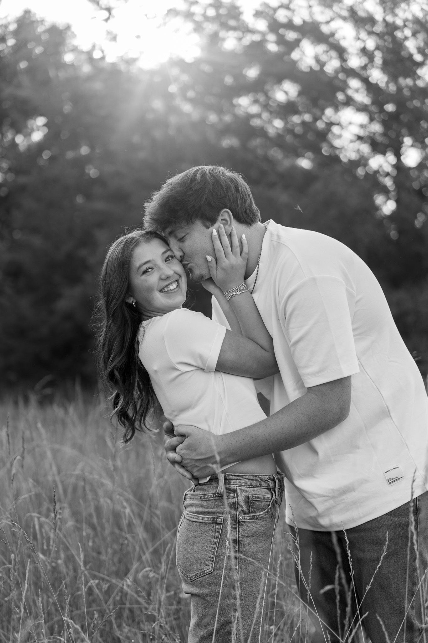 Black and white photo of smiling woman being kissed on cheek by partner in tall grass field.