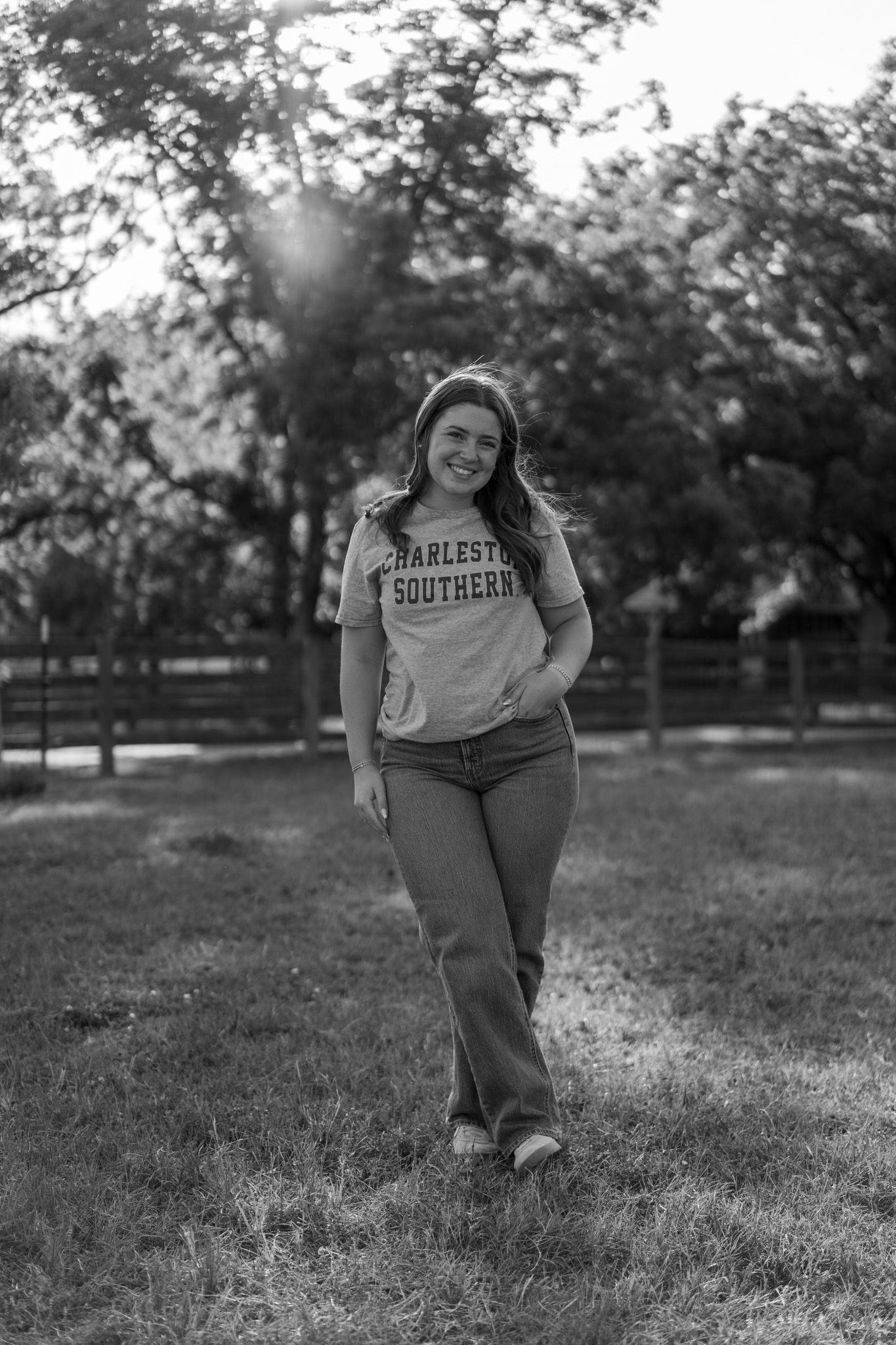 Black and white photo of smiling young woman in Charleston Southern t-shirt walking in sunny park.