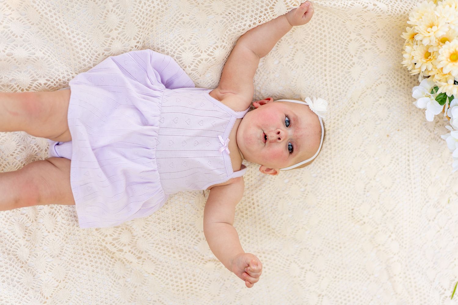 Adorable baby in lavender romper and white bow headband lying on cream crochet blanket with flowers.