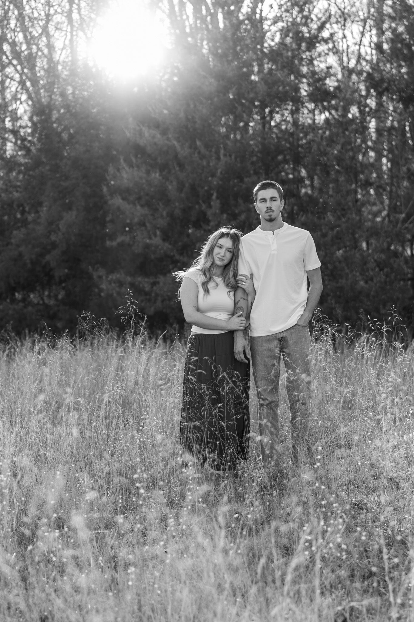 Black and white portrait of couple standing together in tall grass field at sunset.